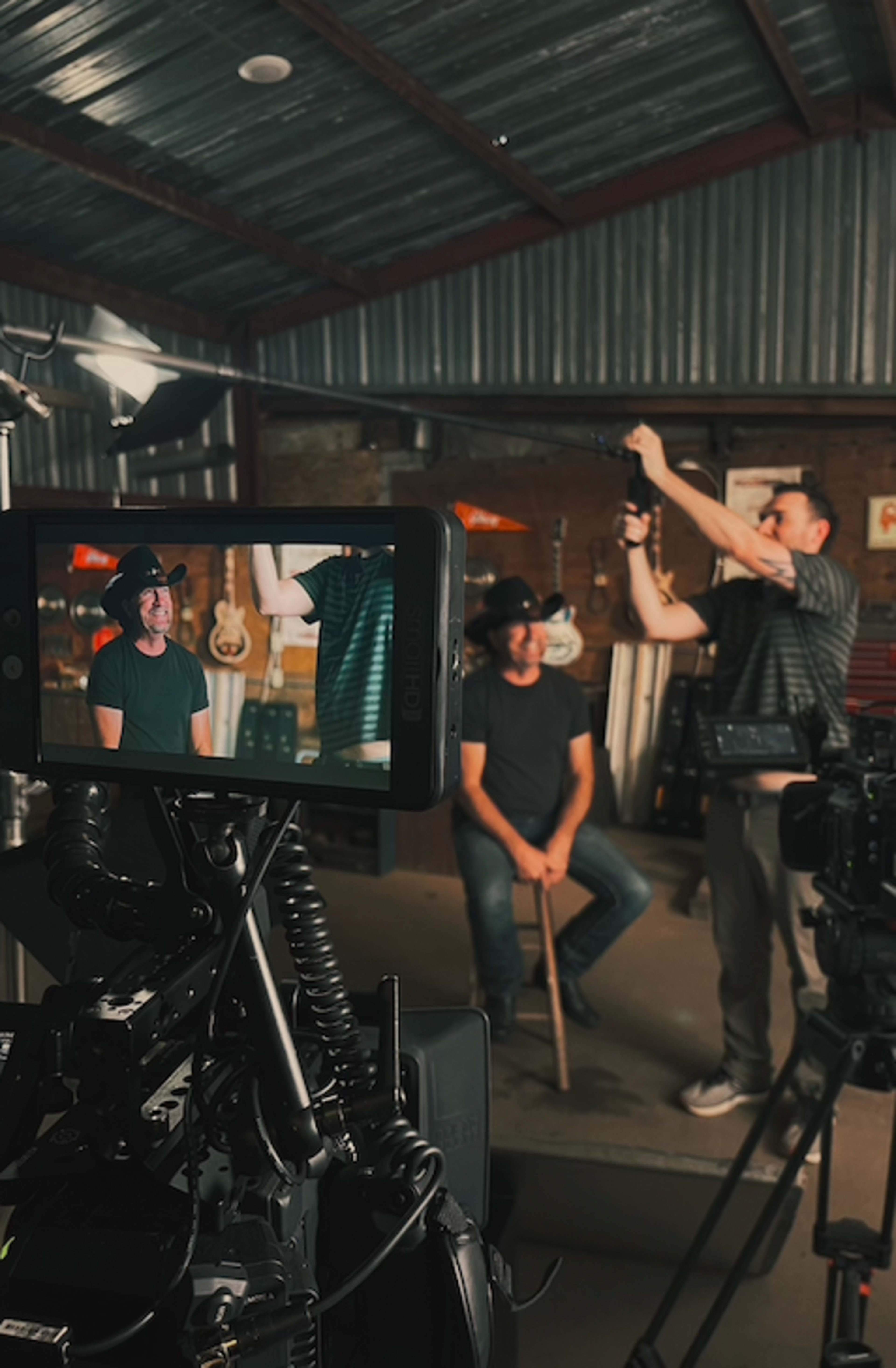 A camera crew films an interview with a seated man holding a cane in a rustic studio filled with guitars.