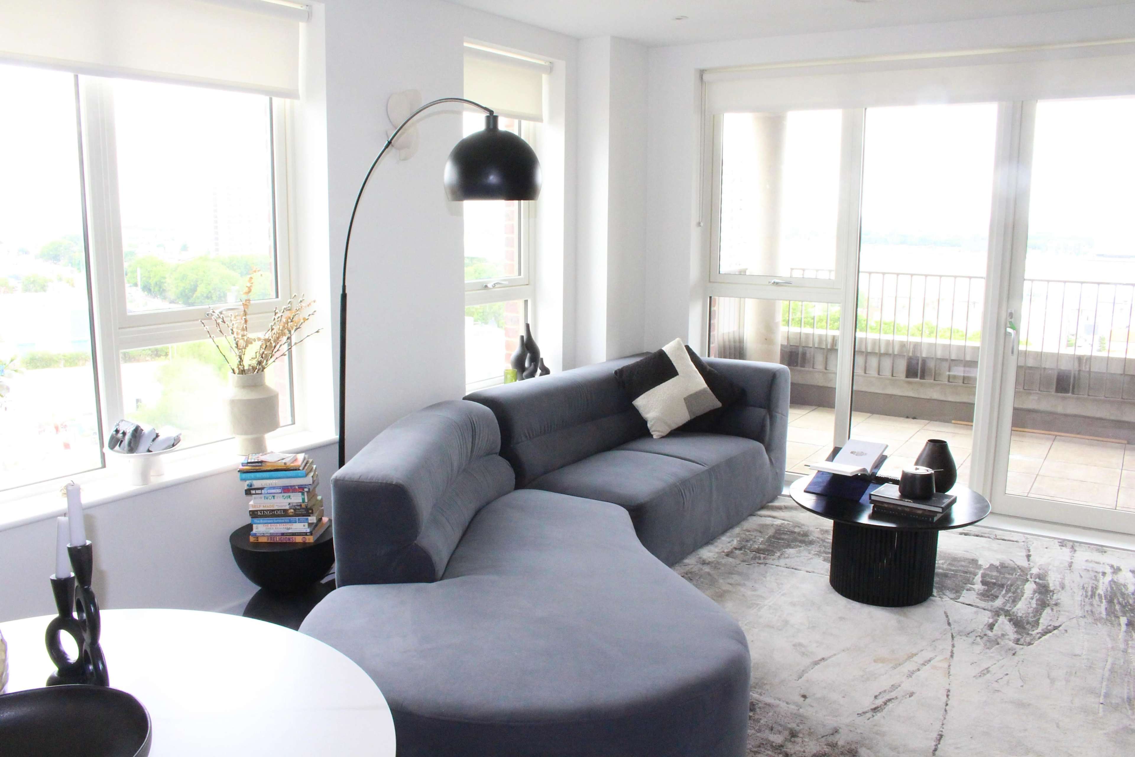 A modern living room features a curved gray sofa, a side table with books, and large windows that provide a view of the outside.