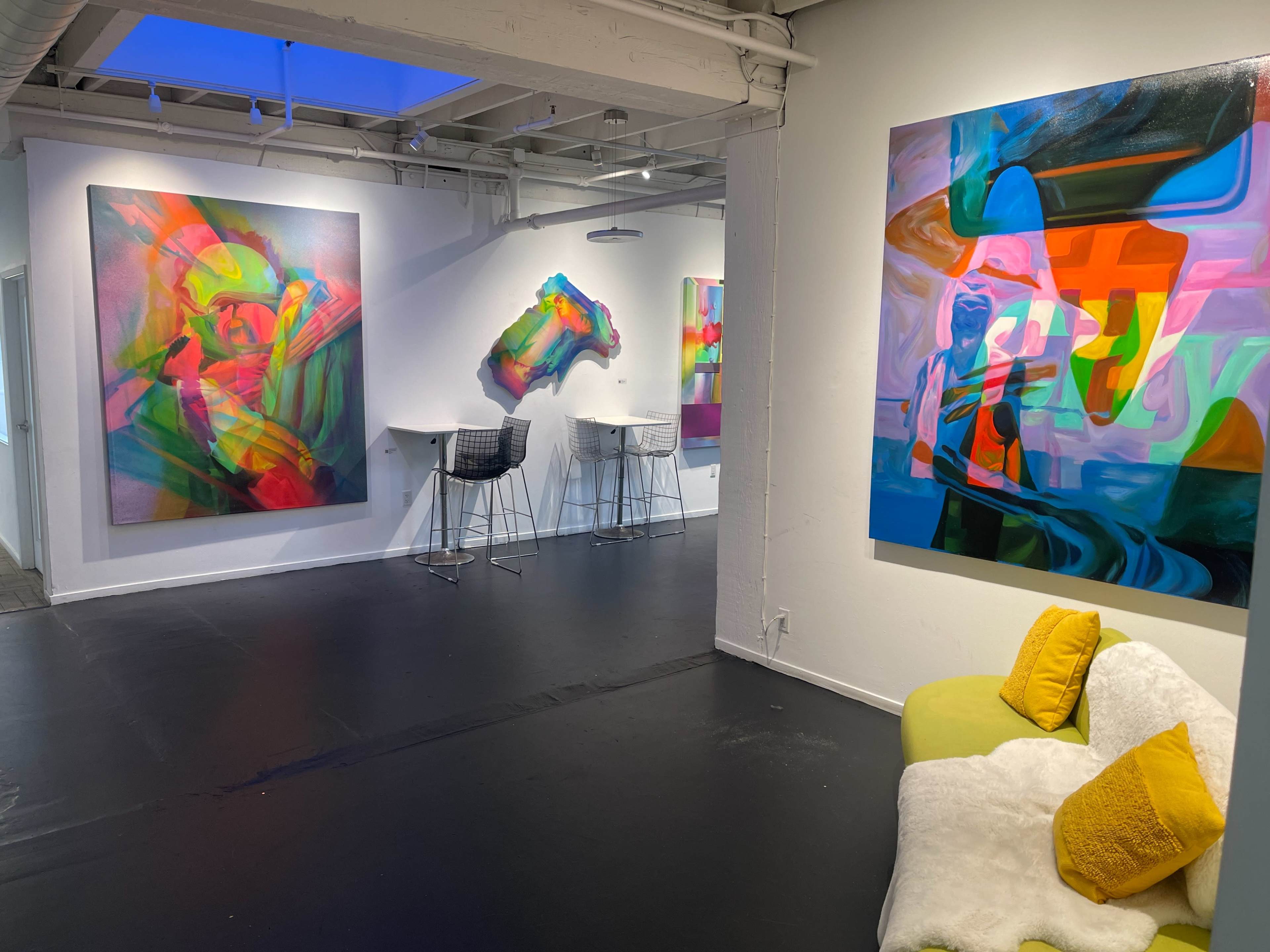 An art gallery with colorful abstract paintings on the walls, bar stools set up along one side, and a couch with decorative pillows in the foreground.