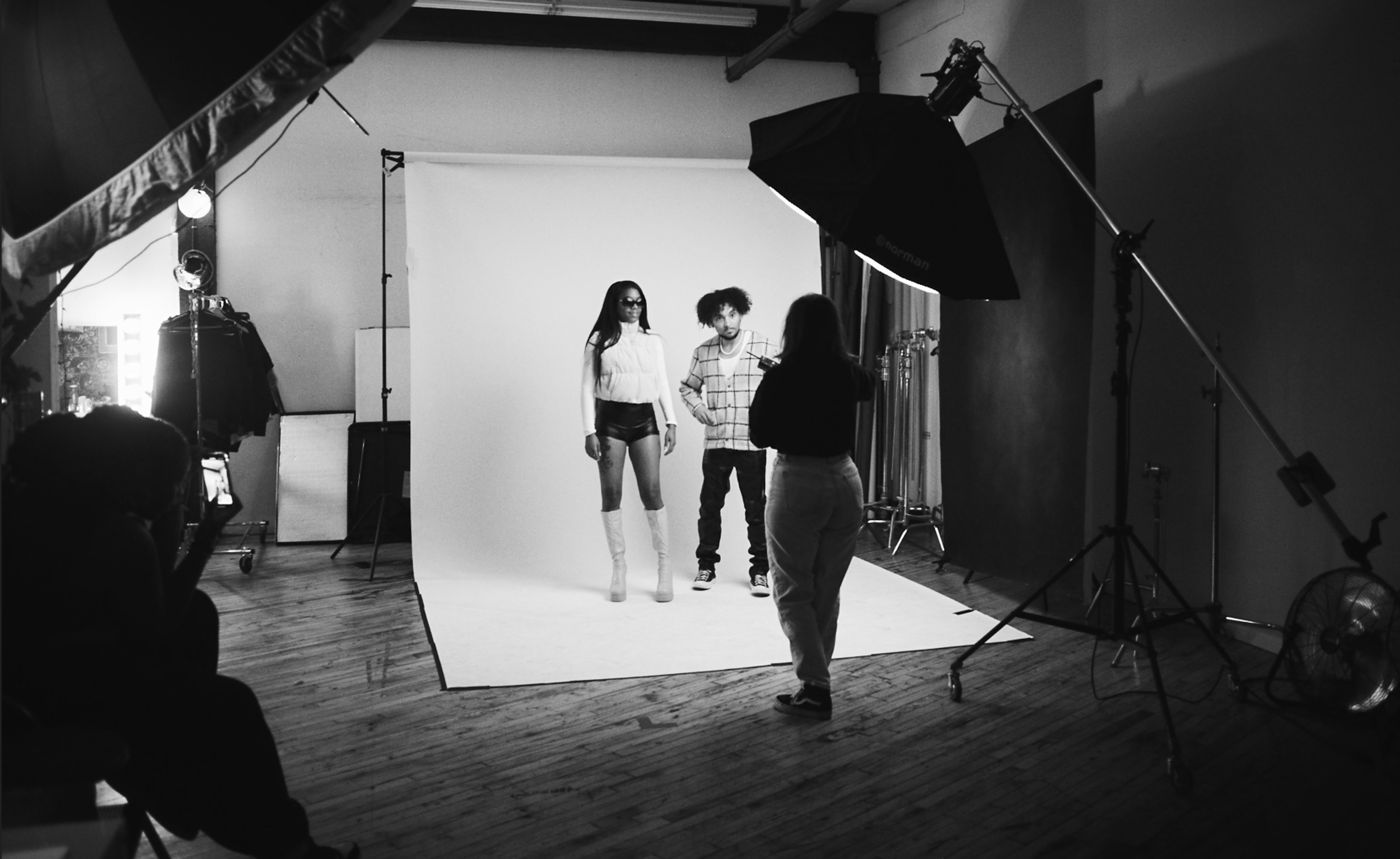 A photoshoot captures two models posing against a plain white backdrop while a photographer directs them.