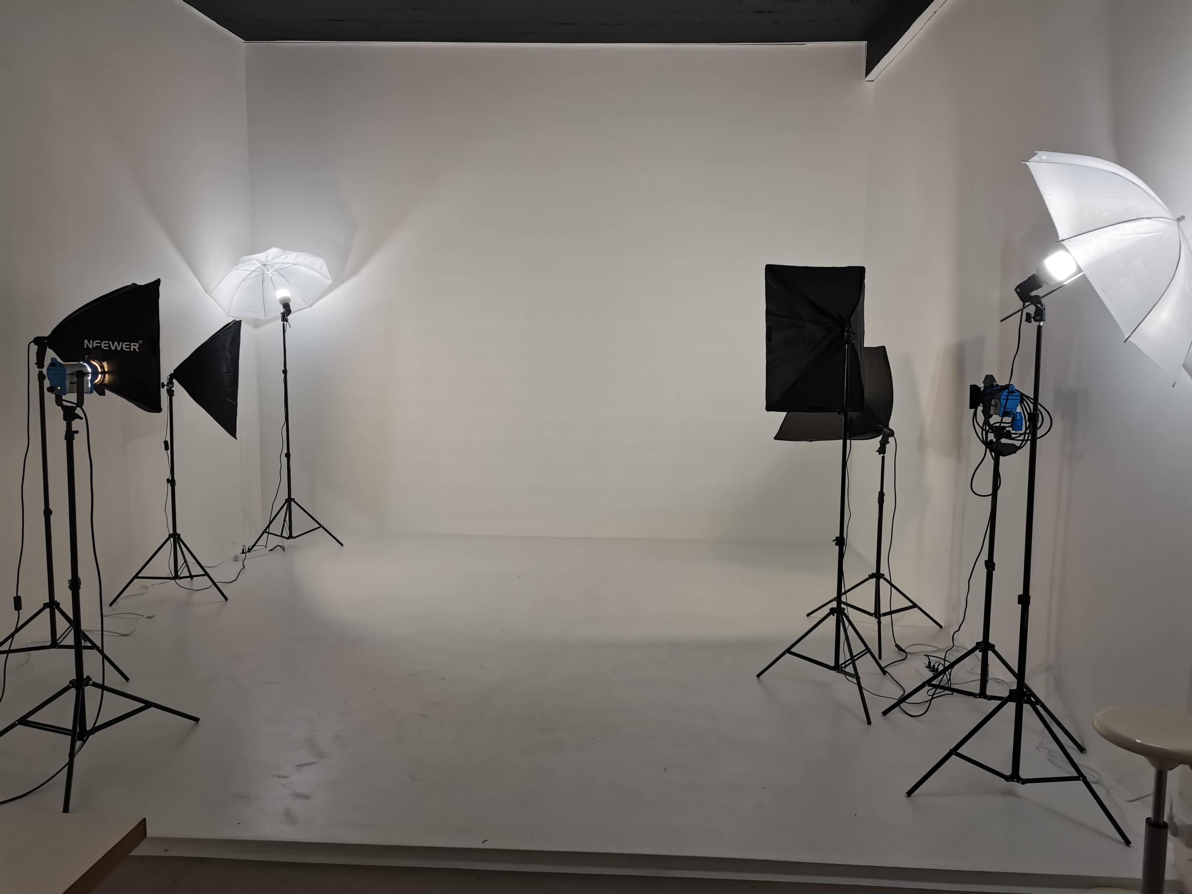 A photography studio is set up with multiple softbox and umbrella lights on tripods, illuminating a plain white wall.
