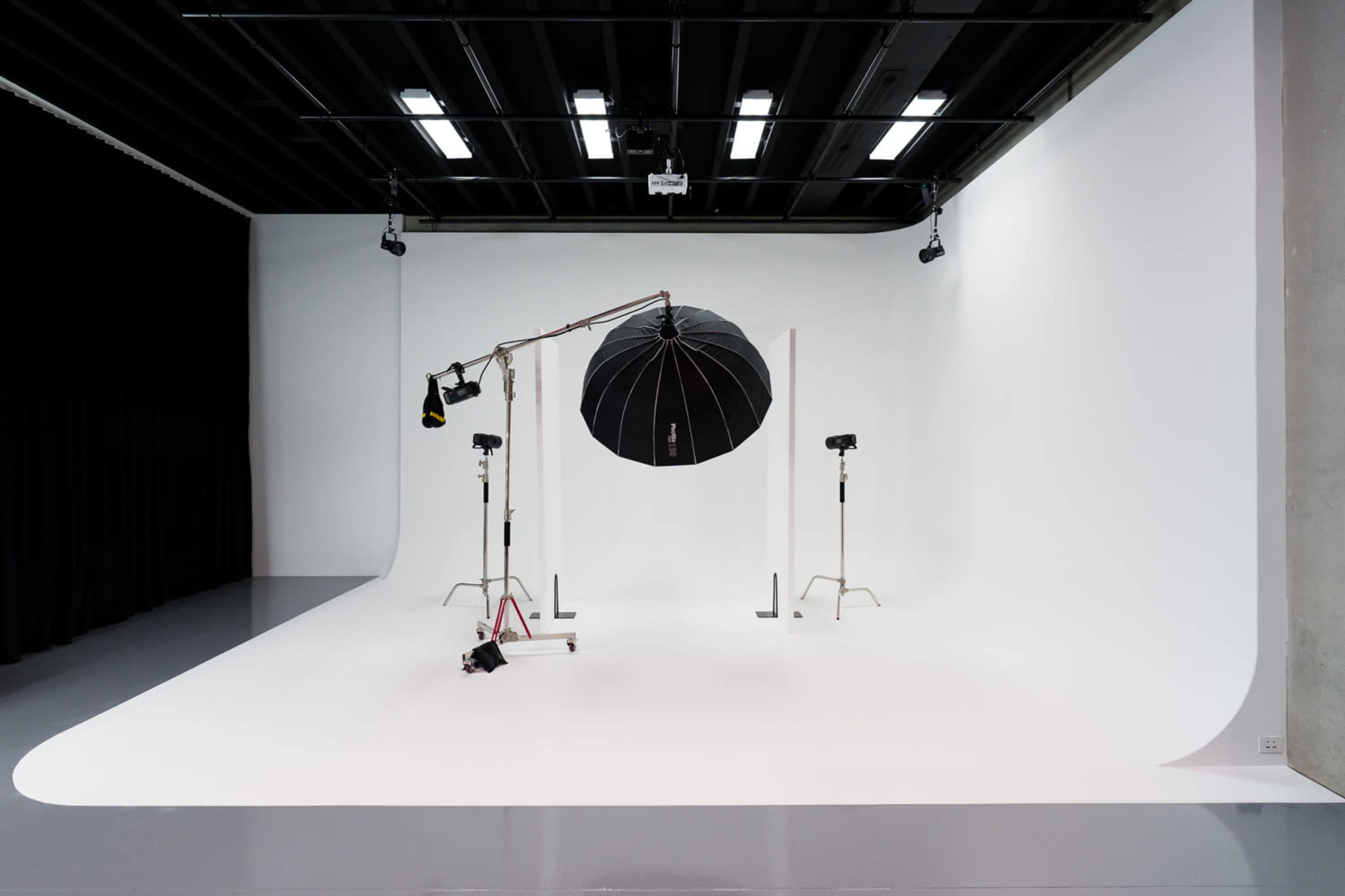 The image shows a photography studio featuring a seamless white backdrop, lighting equipment, and an umbrella reflector suspended from a stand.