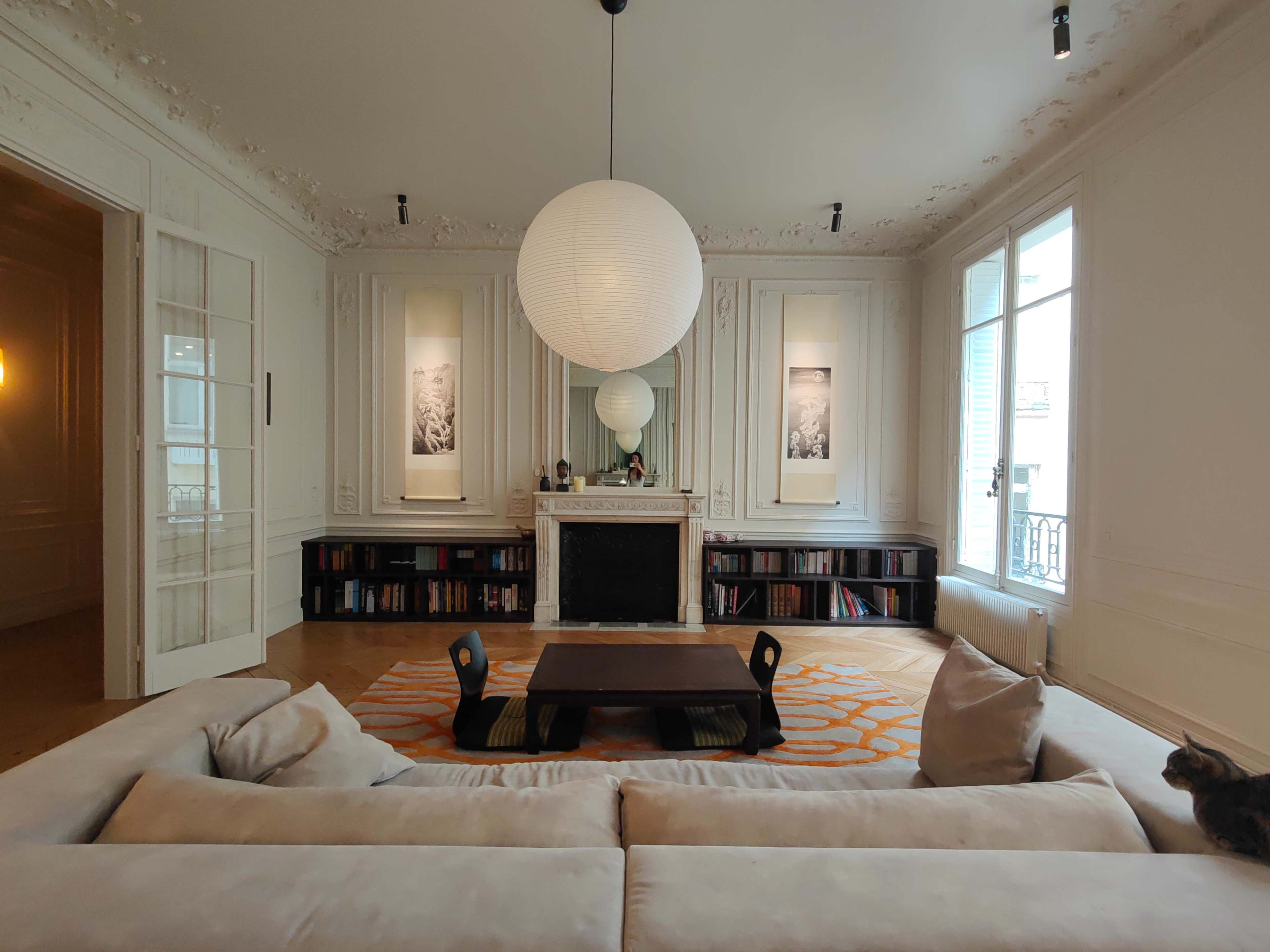 The room features a spacious layout with a large sofa, a central coffee table, a fireplace, and shelves filled with books, all illuminated by a large pendant lamp.