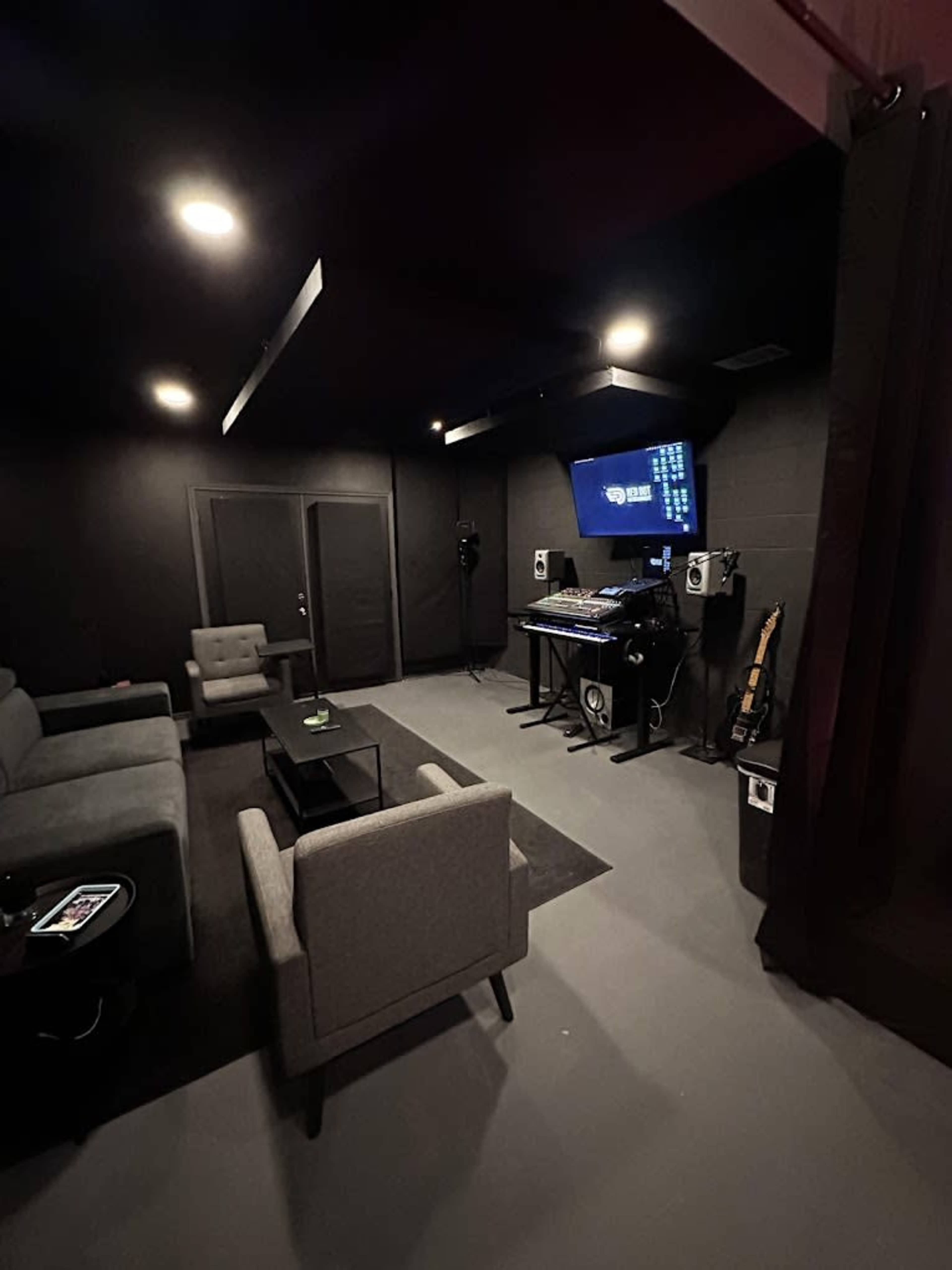 The image shows a dimly lit room featuring a seating area with gray furniture, a glass coffee table, and a music equipment setup against one wall.