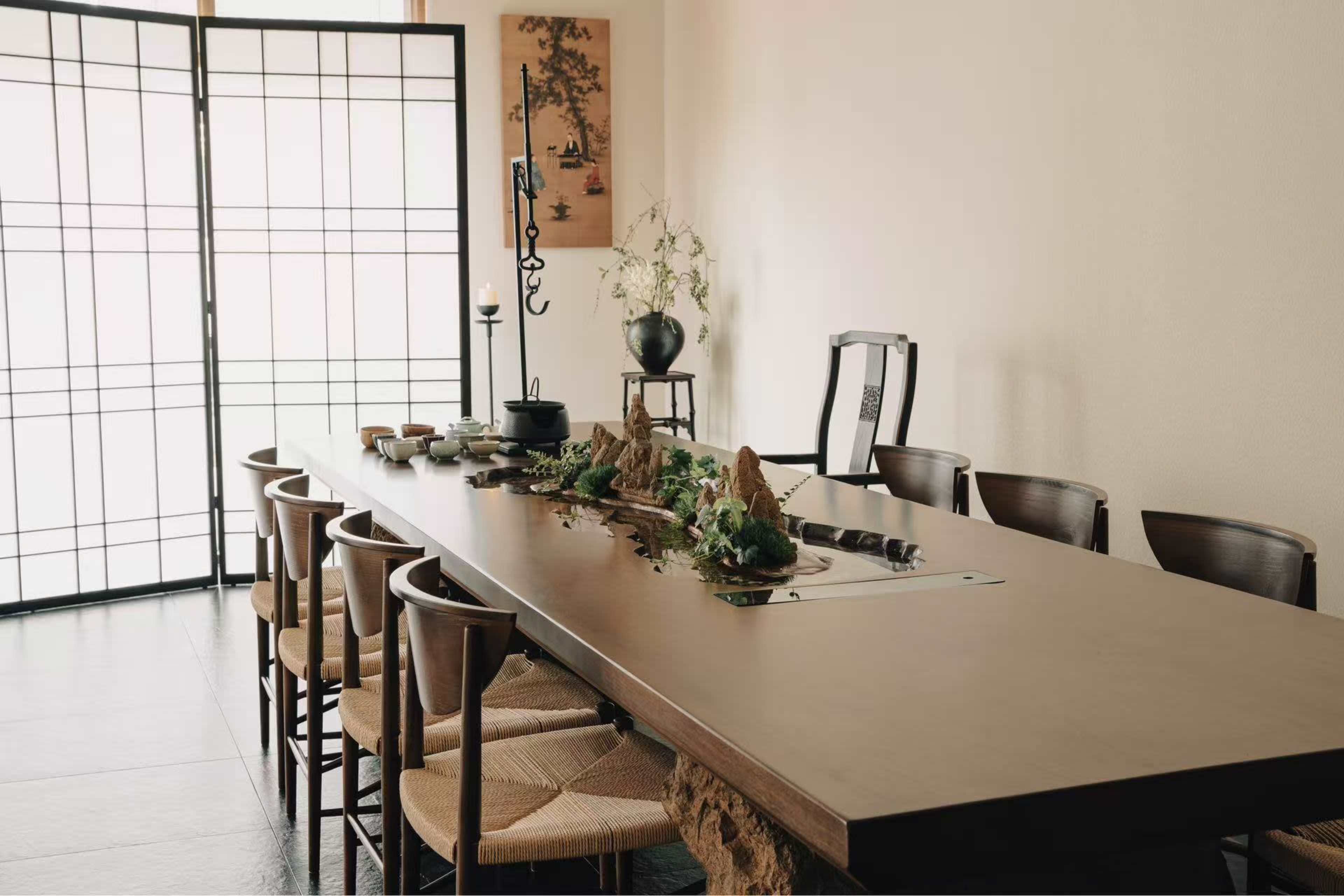 The image shows a minimalist dining room featuring a long table set with decorative elements and surrounded by wooden chairs, with a backdrop of sliding shoji screens.