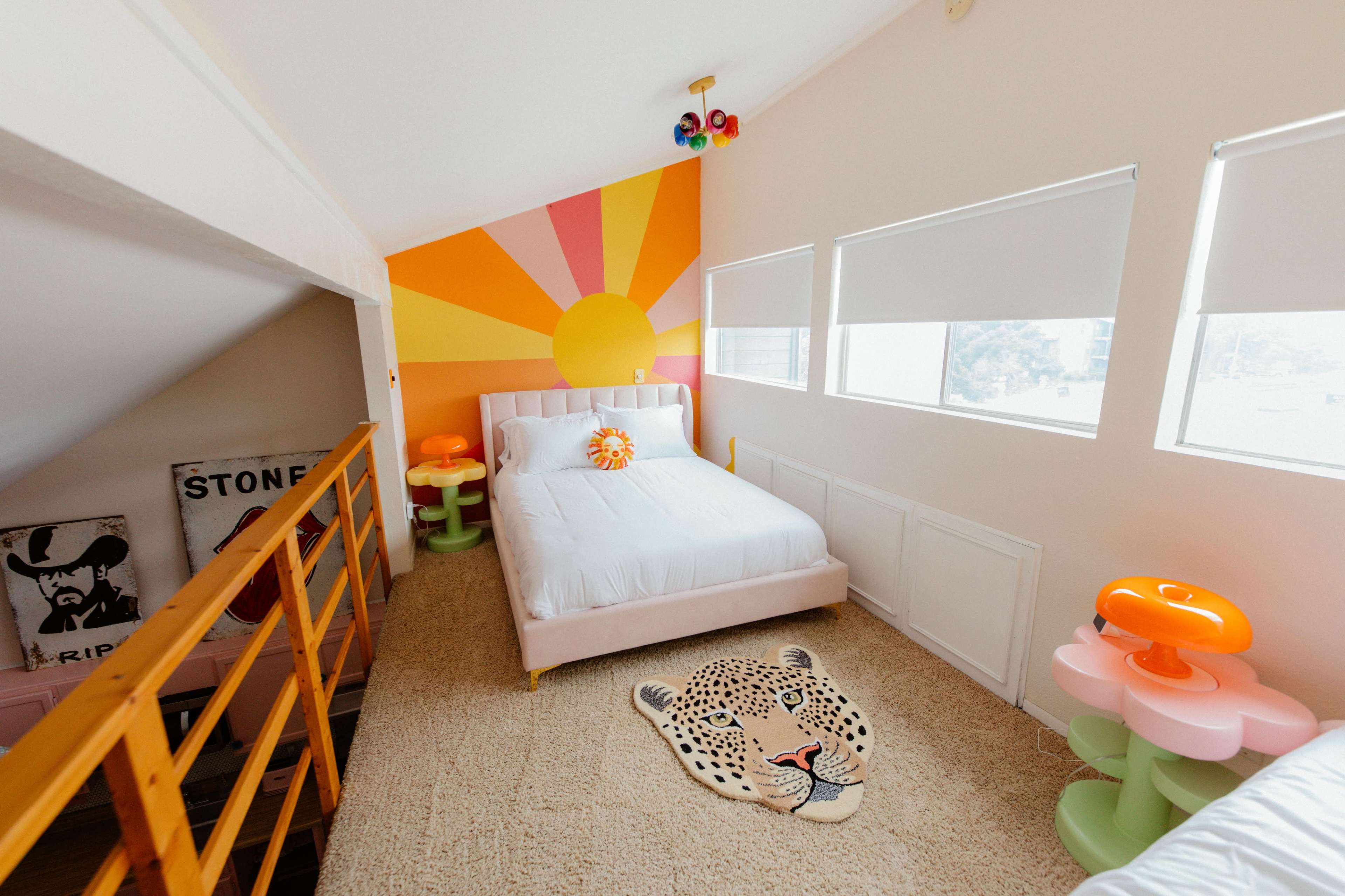 The image shows a brightly colored bedroom with a sun mural on the wall, a bed with a light pink headboard, and a leopard-print area rug on a carpeted floor.