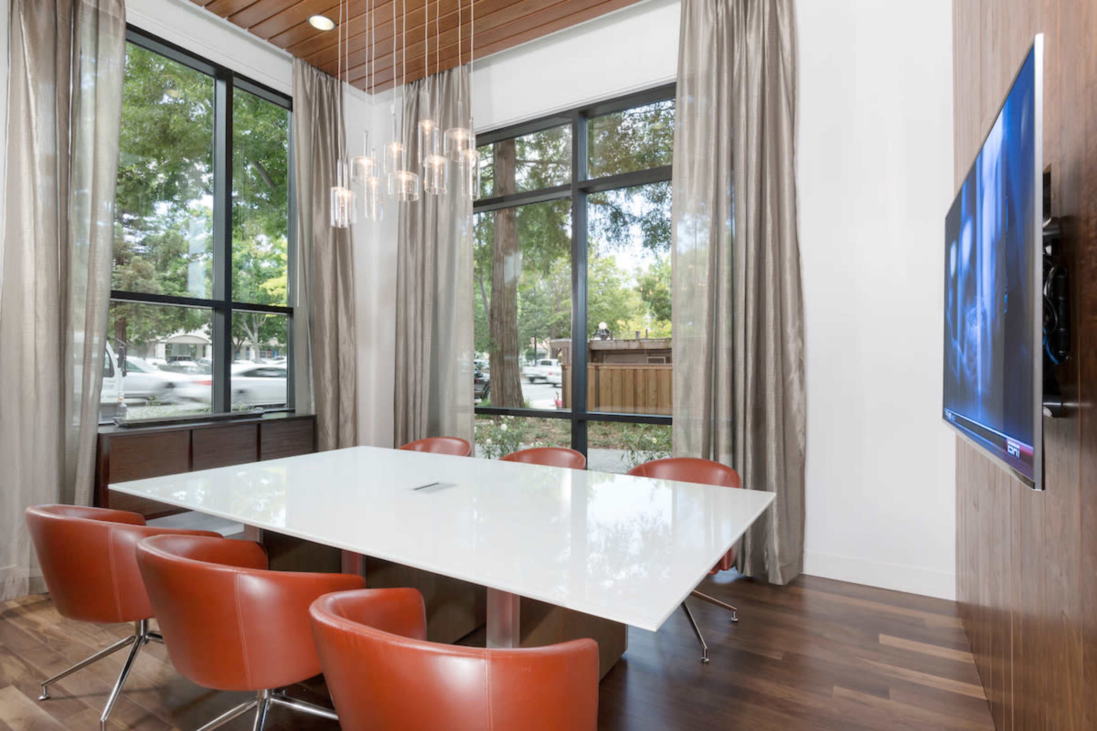 A modern dining area features a sleek white table surrounded by orange chairs, with large windows allowing natural light and a view of trees outside.
