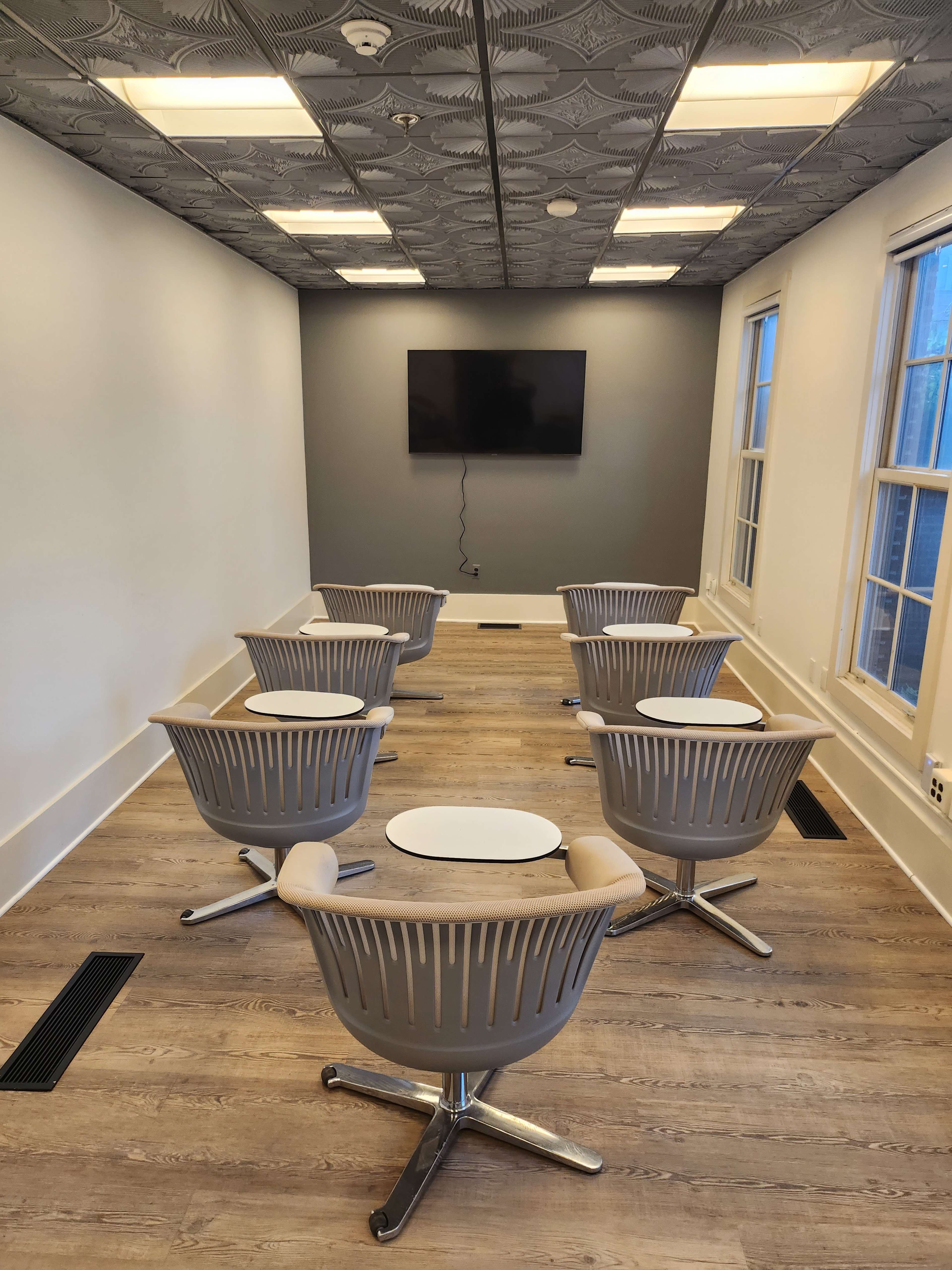 A modern meeting room features six gray chairs arranged around a small round table, facing a wall-mounted television.