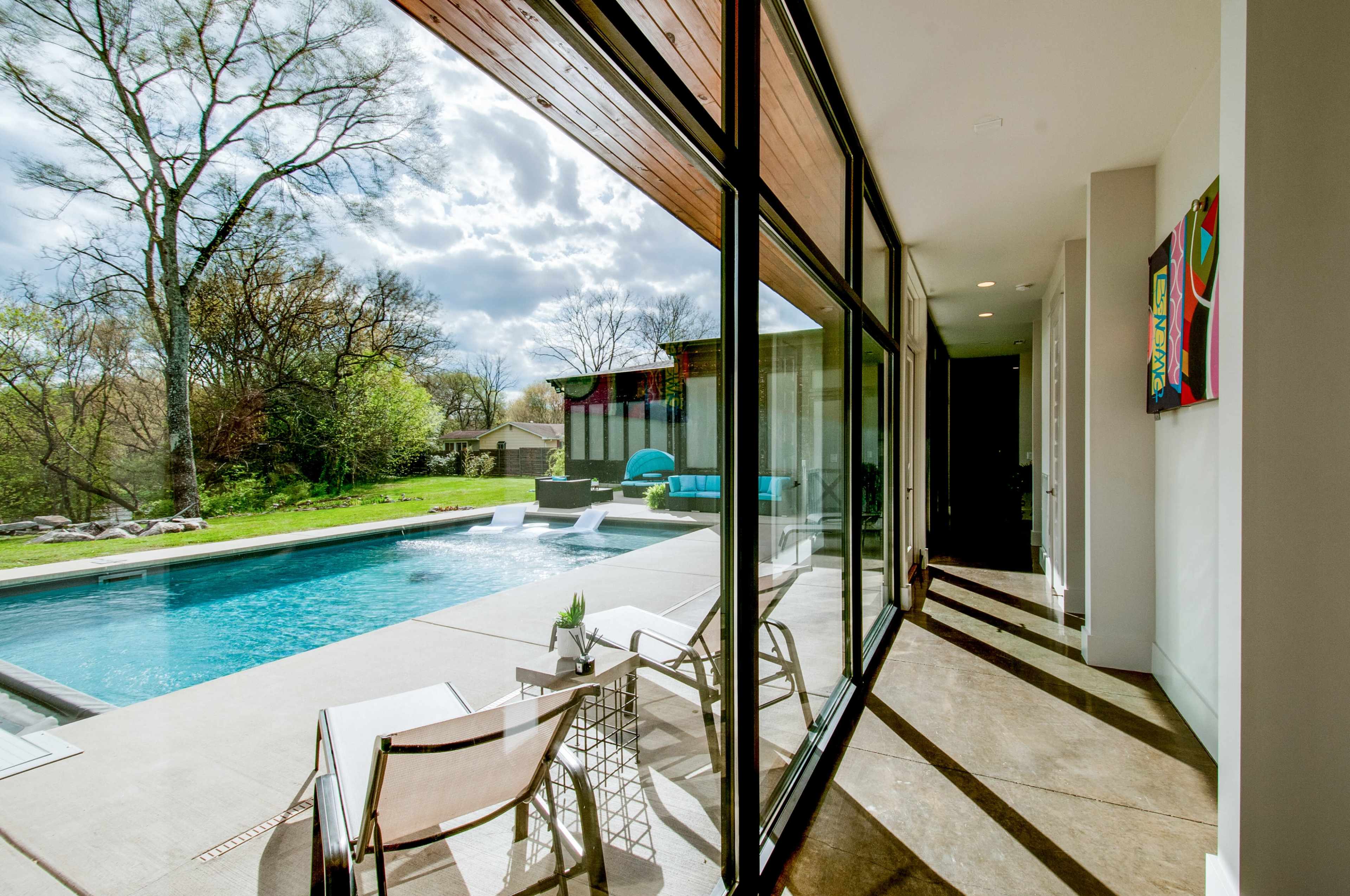 A modern home features large glass doors opening to a poolside area with patio chairs, surrounded by greenery and clouds in the sky.
