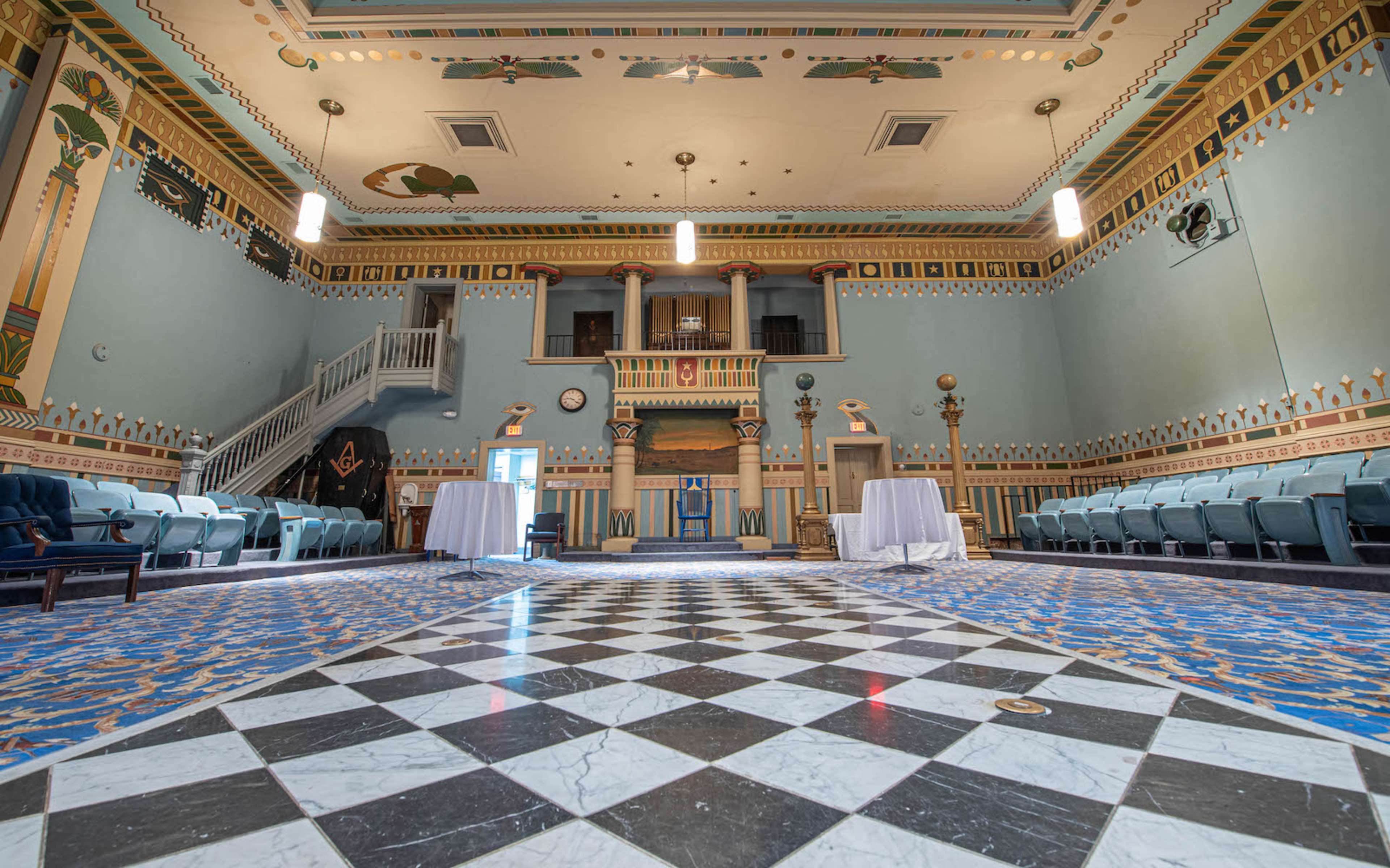 A spacious, ornately decorated room with a checkered floor, featuring chairs arranged in rows and tables set up in the center.