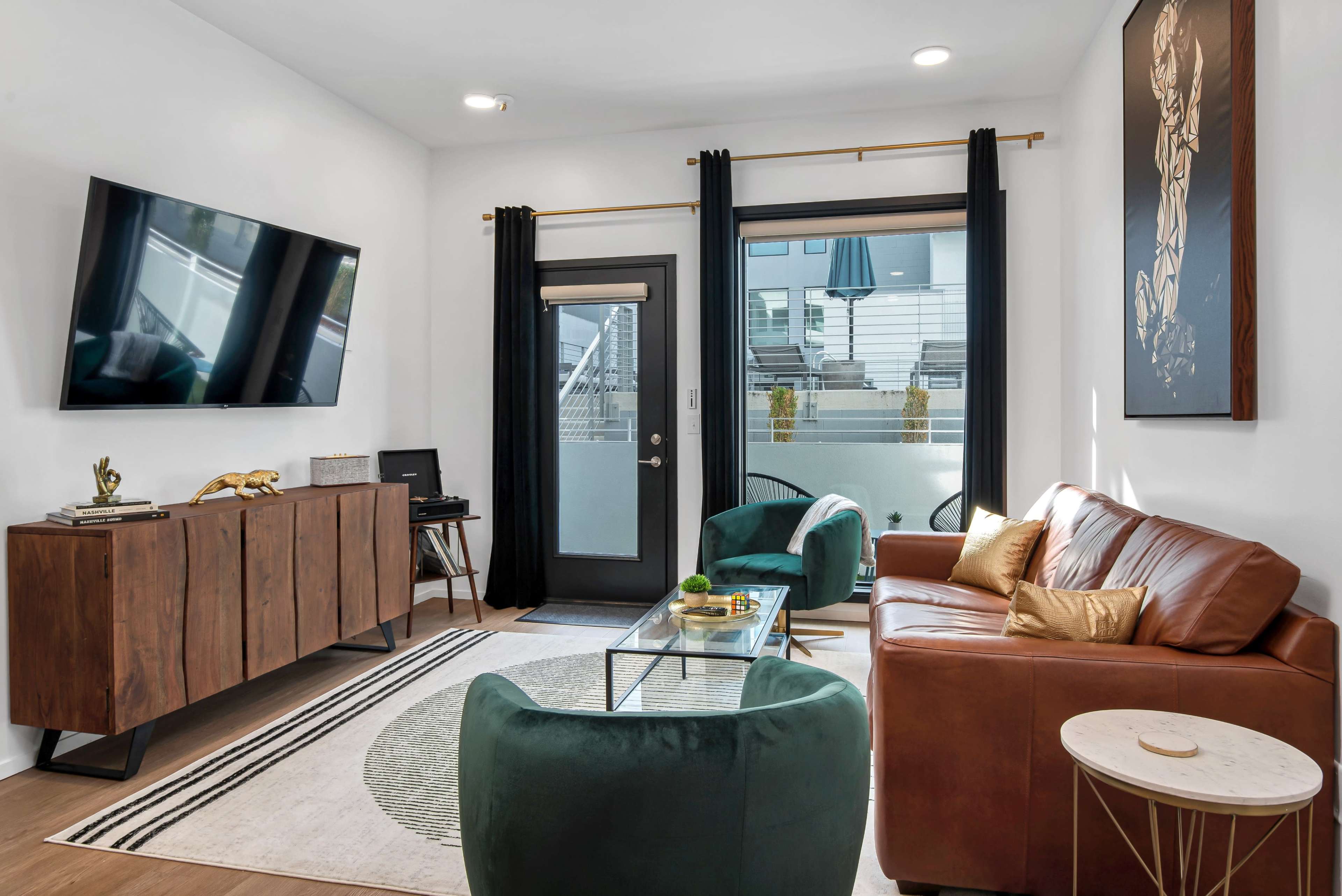 The living room features a brown leather sofa, green accent chairs, a glass coffee table, and a large wall-mounted TV, with floor-to-ceiling windows and black curtains.
