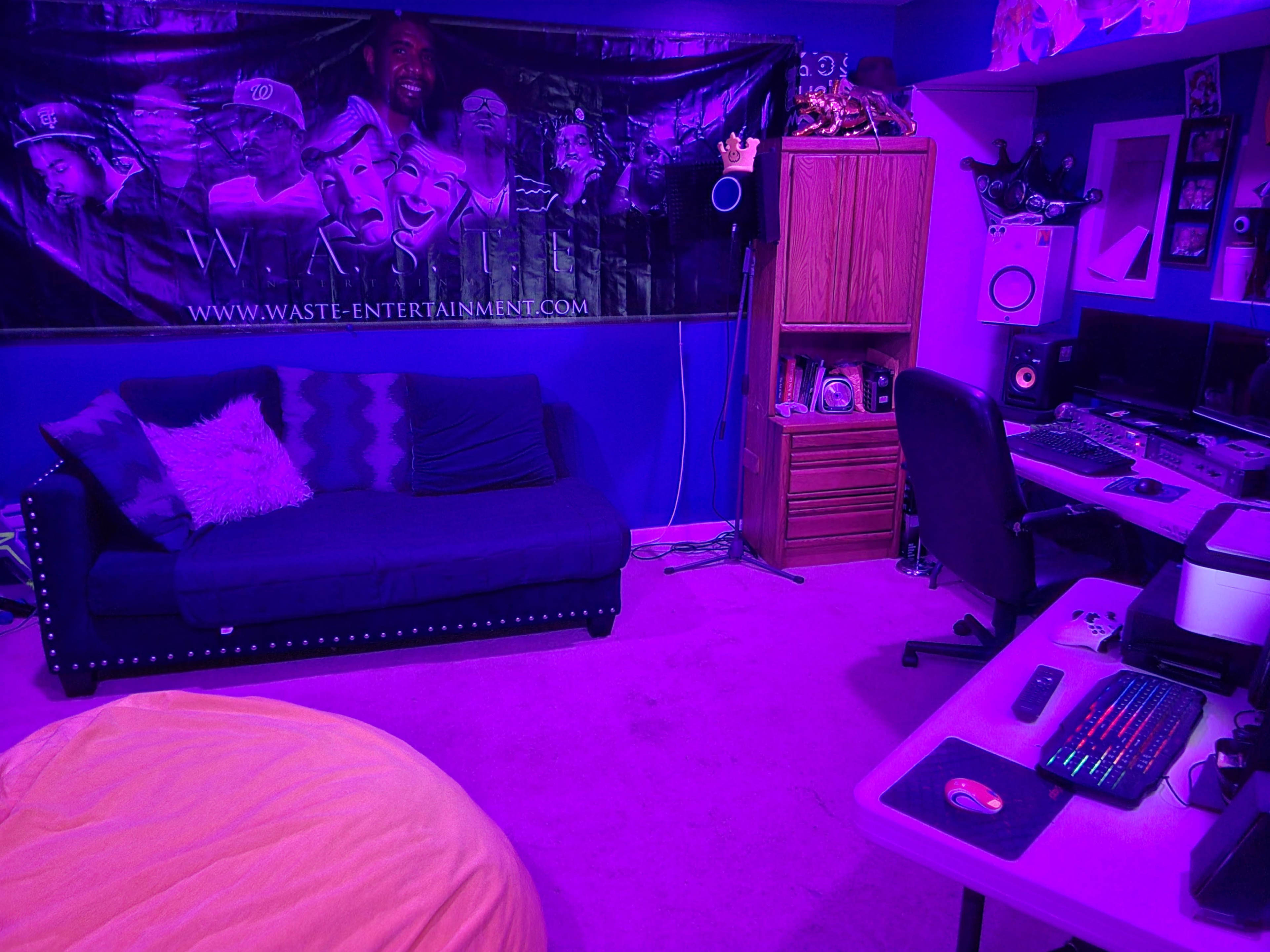 A small room featuring a purple-lit couch, a desk with computer equipment, and a large decorative wall banner.