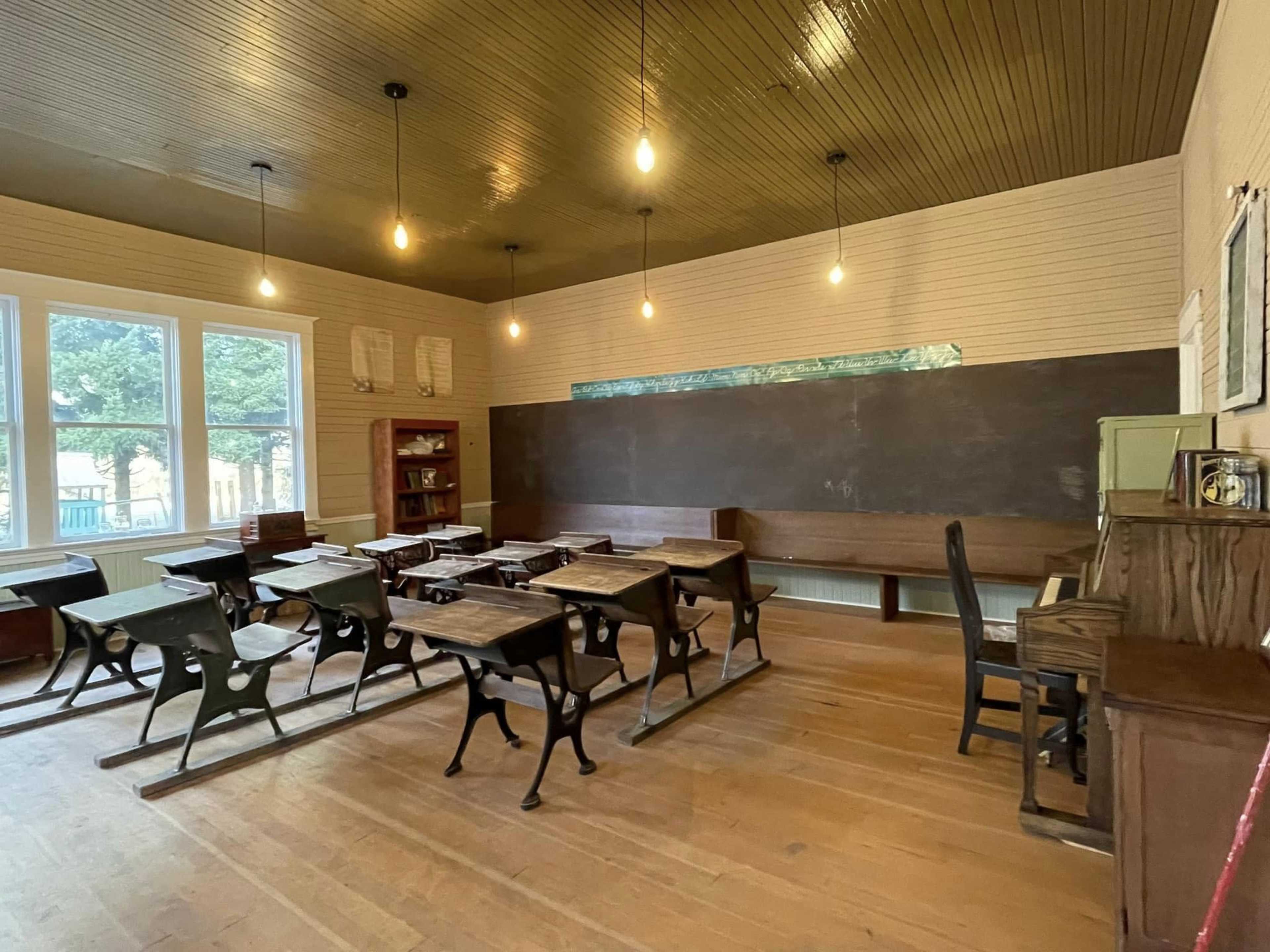 A vintage classroom contains wooden desks, a chalkboard, and a bookshelf, illuminated by hanging light bulbs.