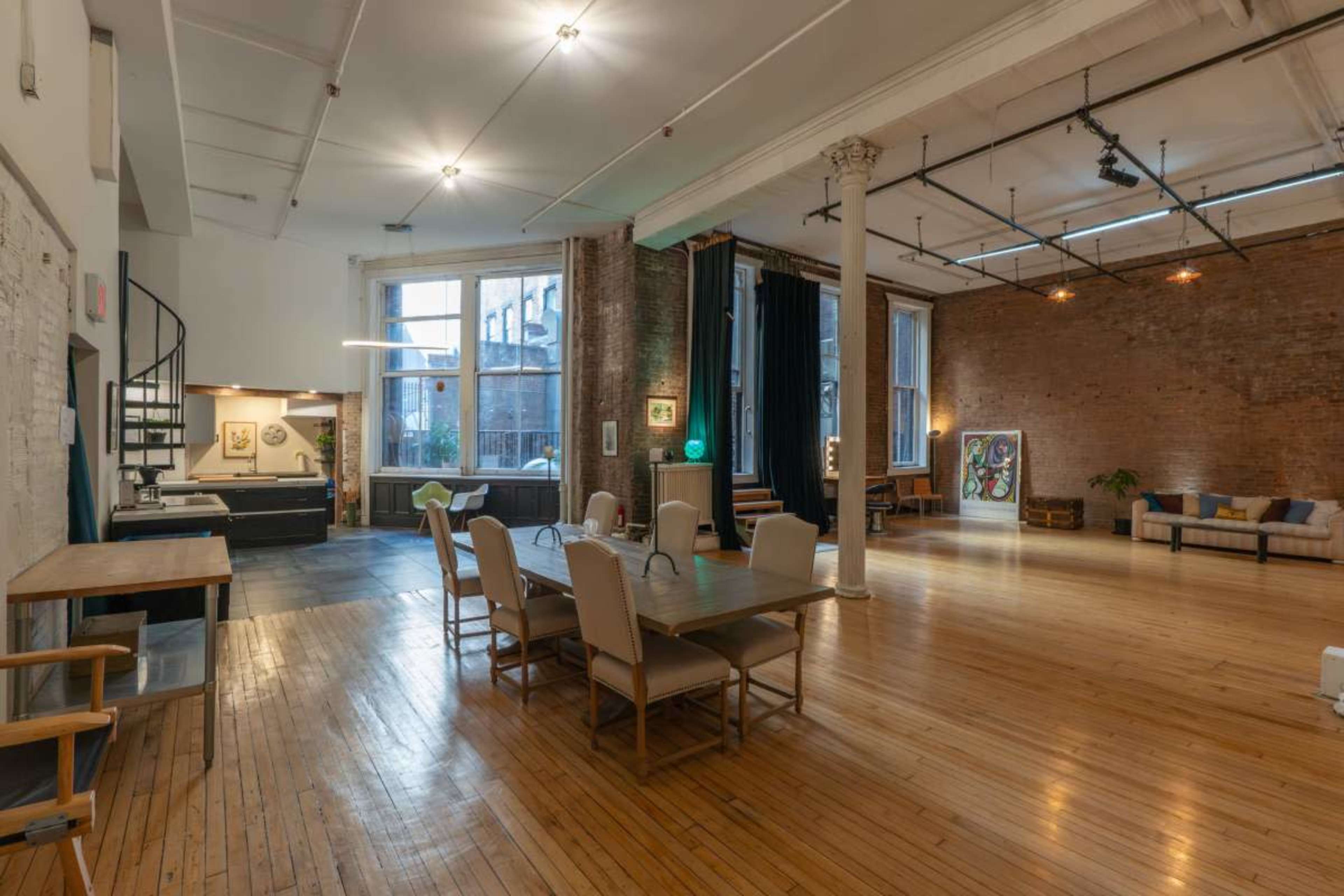 The image shows a spacious, open interior with wooden floors, large windows, a dining table with chairs, and a seating area featuring a sofa and art on the walls.