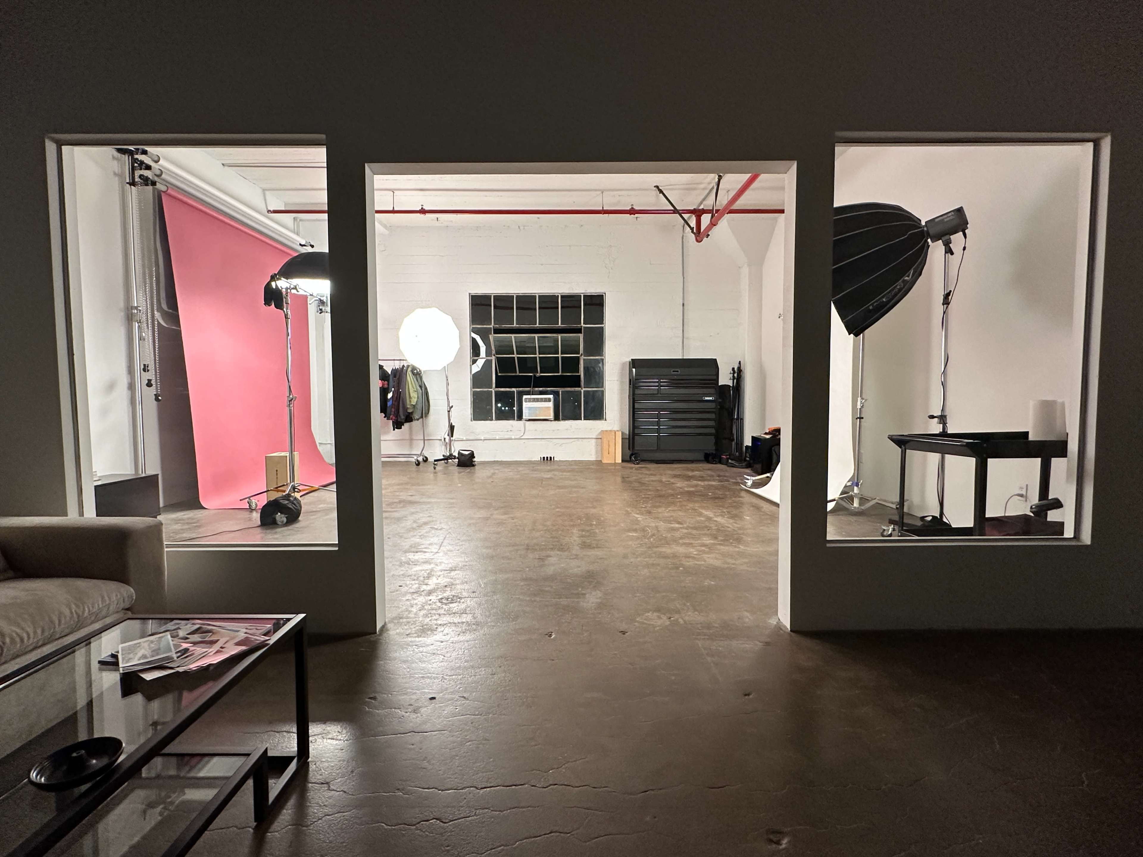 The image shows a spacious studio with three open windows, featuring a pink backdrop, lighting equipment, and polished concrete floors.