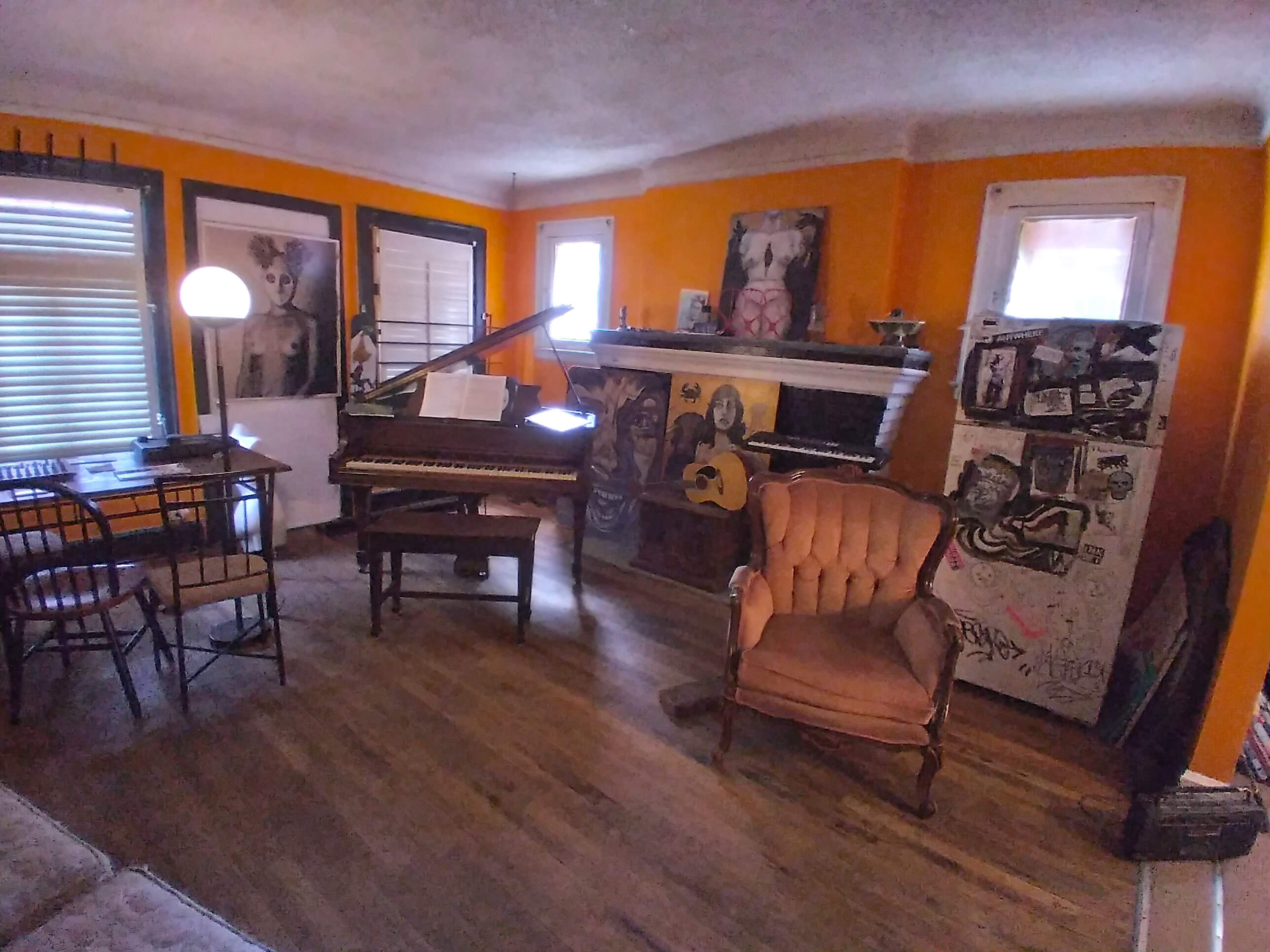 The image shows a colorful room with a piano, a vintage armchair, a dining table with chairs, and eclectic artwork on the walls.
