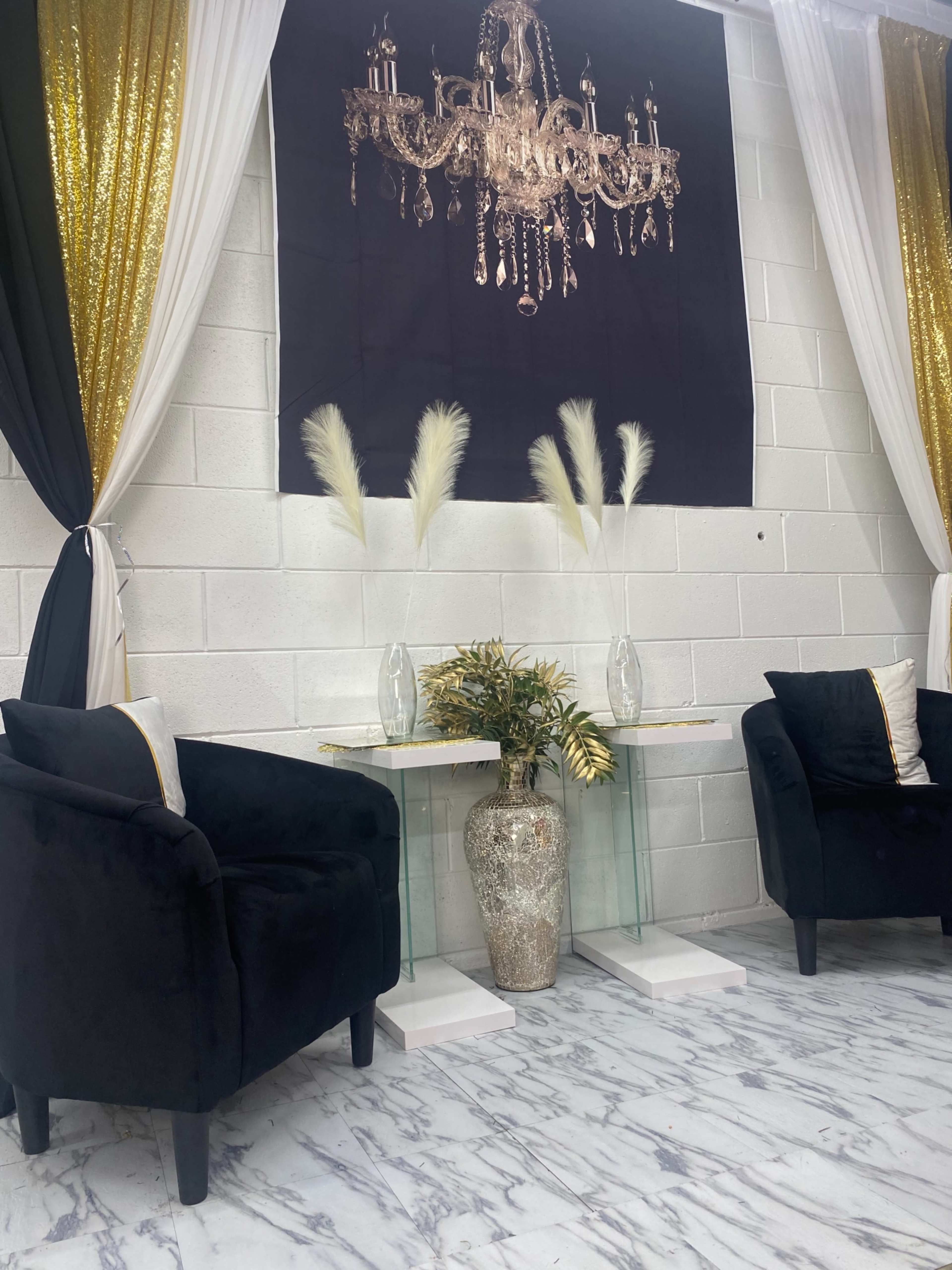 The scene features two black chairs positioned beside a glass table featuring a decorative vase and dried plants, under a chandelier display backdrop with gold and black drapery.