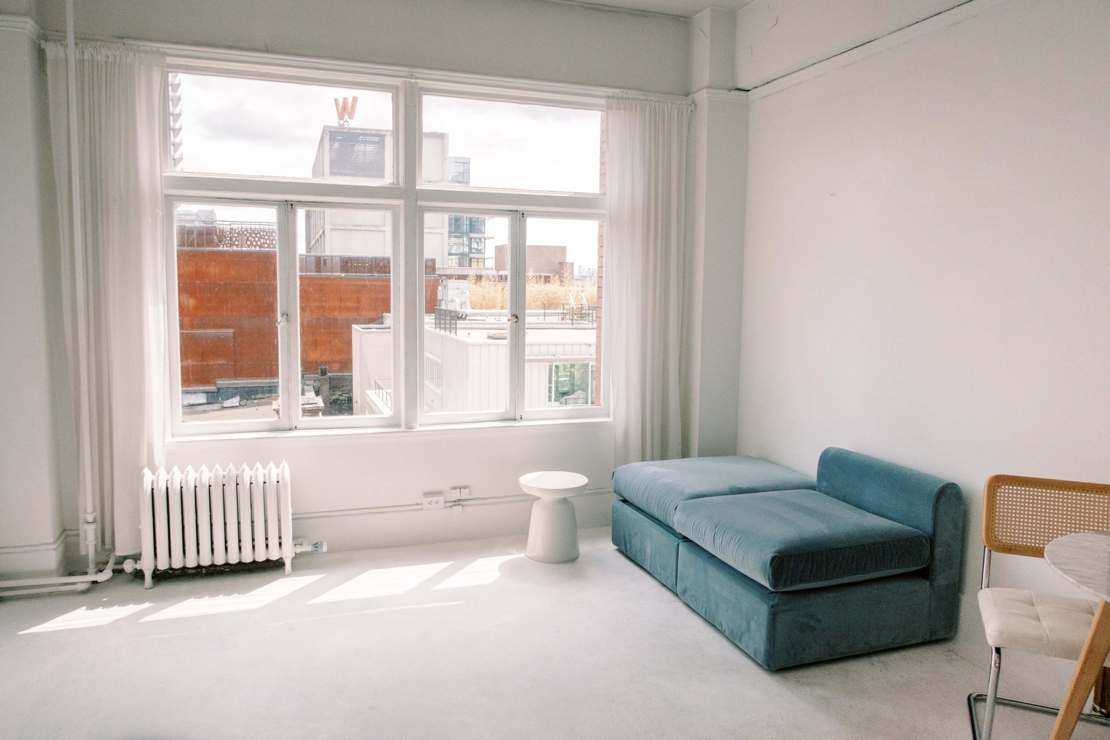 A sunlit room features a blue couch, a small round table, and a view of an urban landscape through large windows.