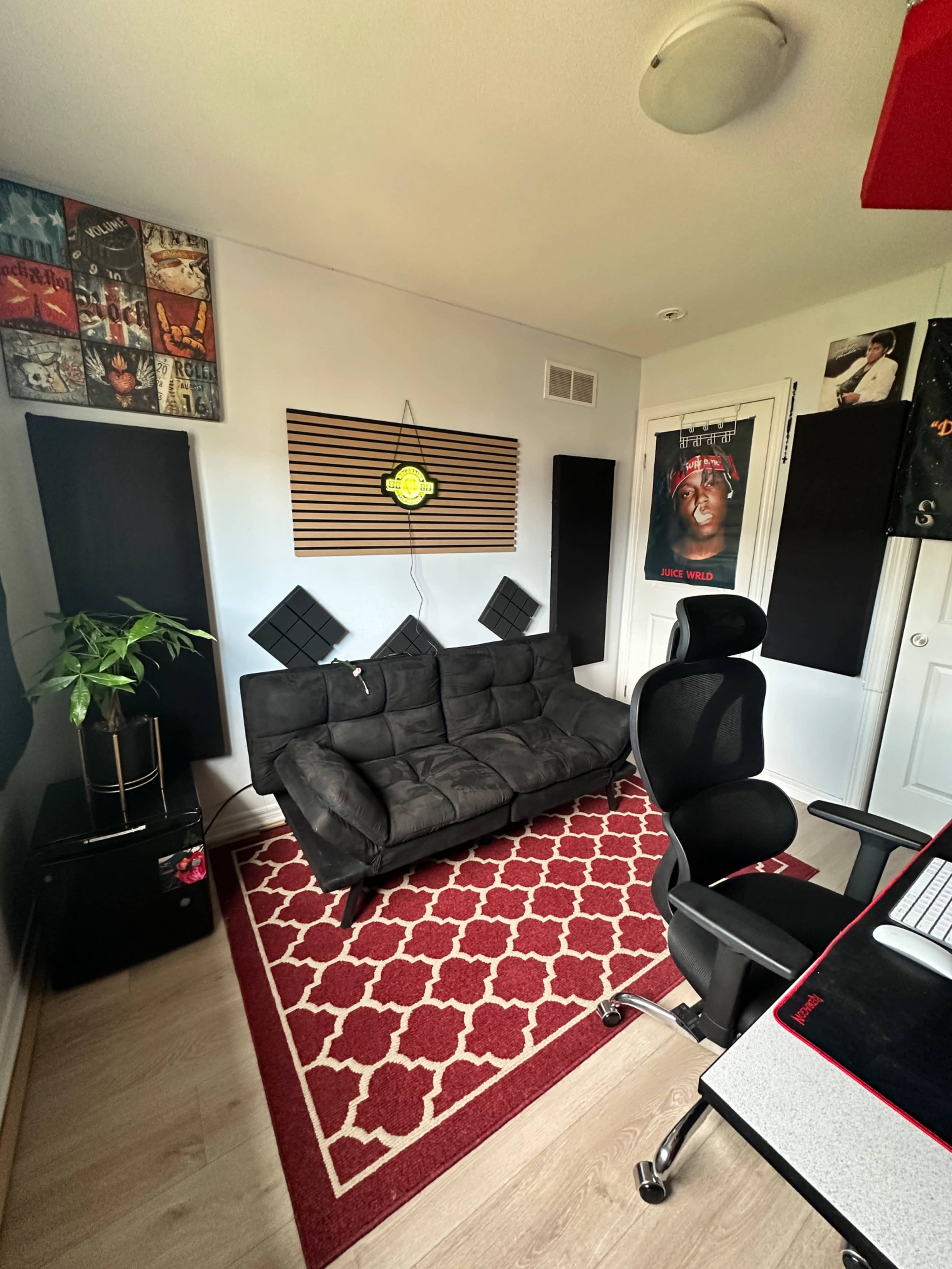 The image shows a small recording studio or office space featuring a black couch, a red patterned rug, soundproofing panels on the walls, a potted plant, and a black office chair near a computer setup.