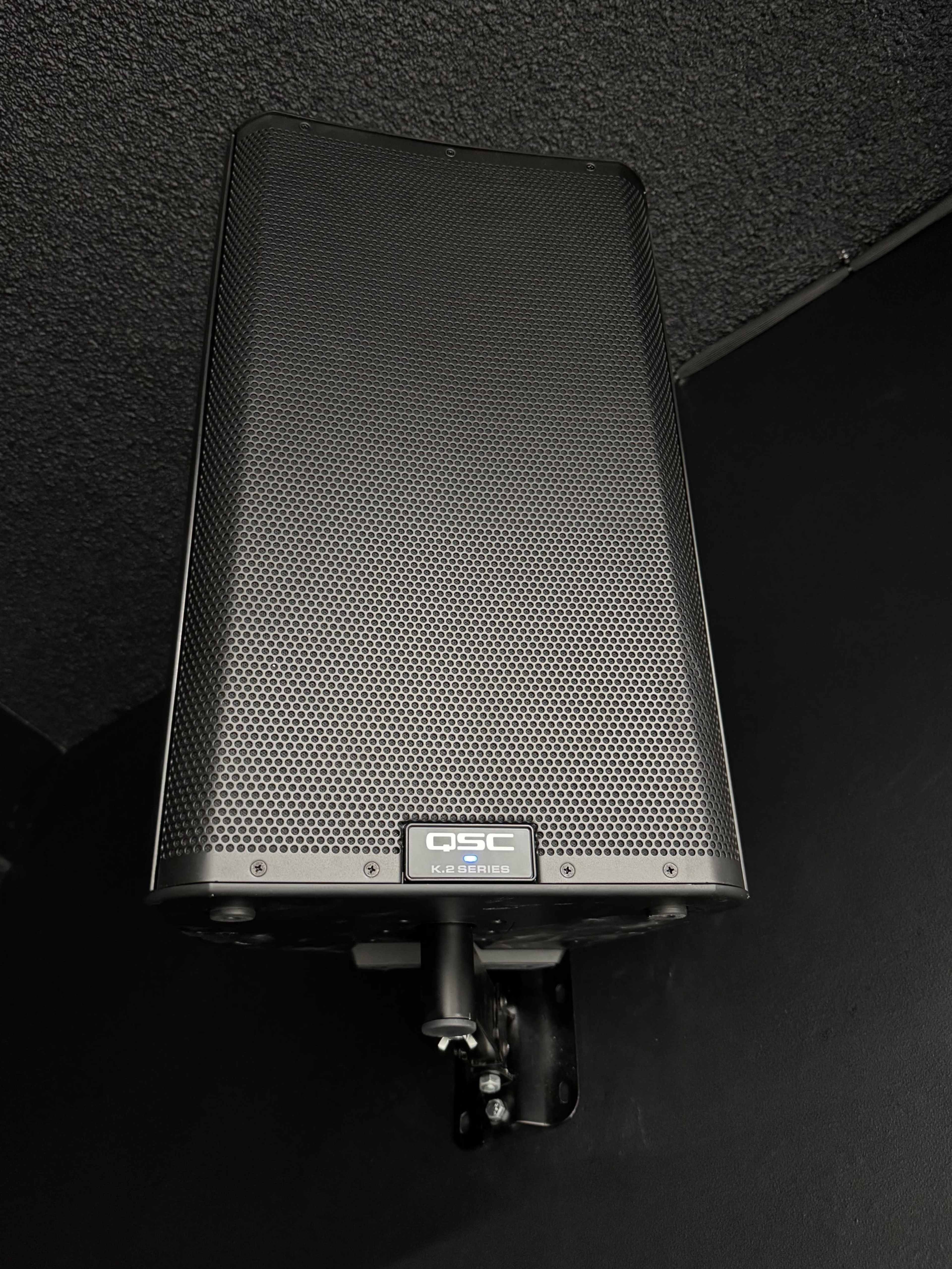 A black QSC K8.2 speaker is mounted on a wall in a dark environment.