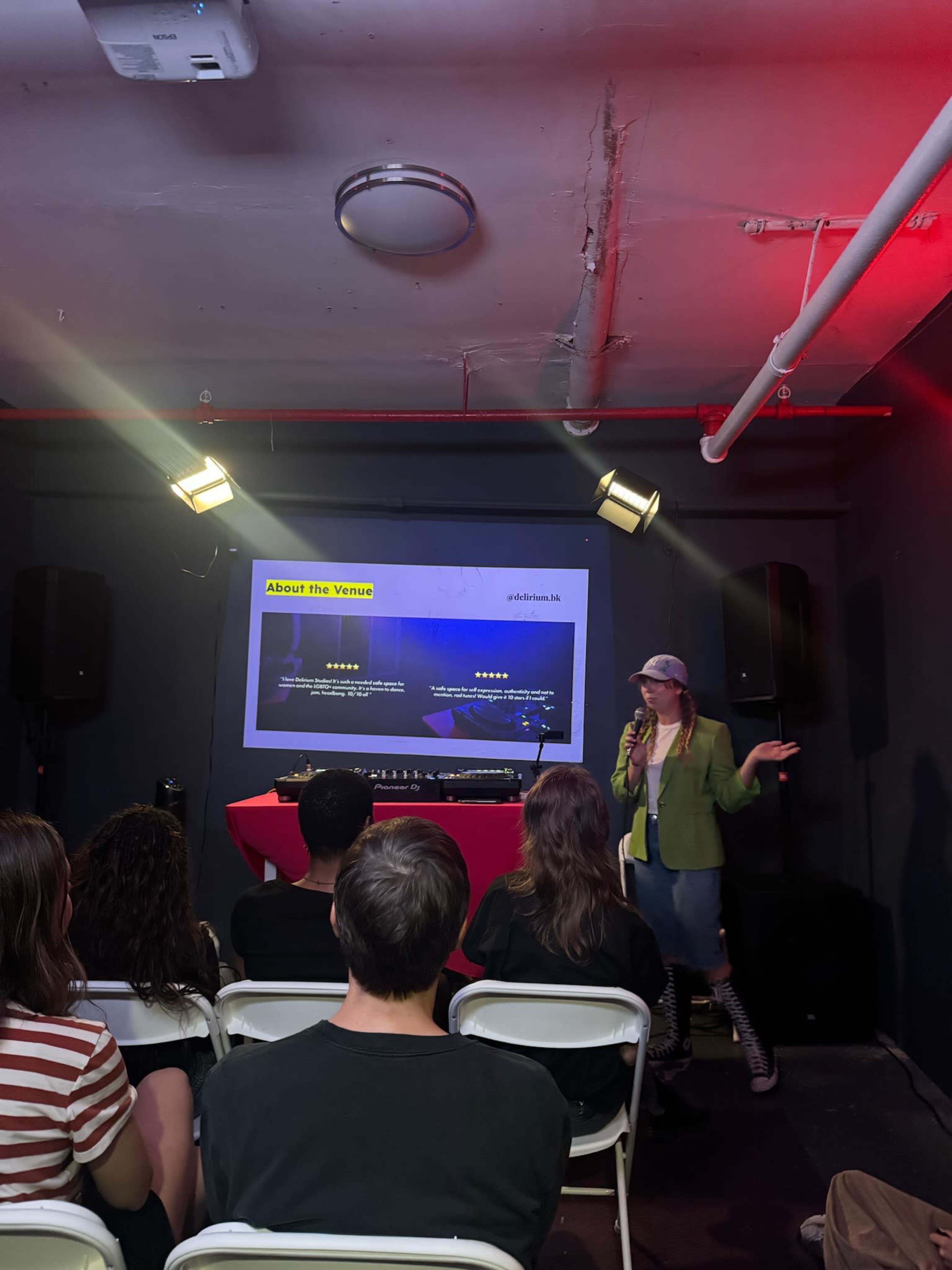 A presenter stands in front of a screen, addressing an audience seated in chairs, with DJ equipment visible on a table.