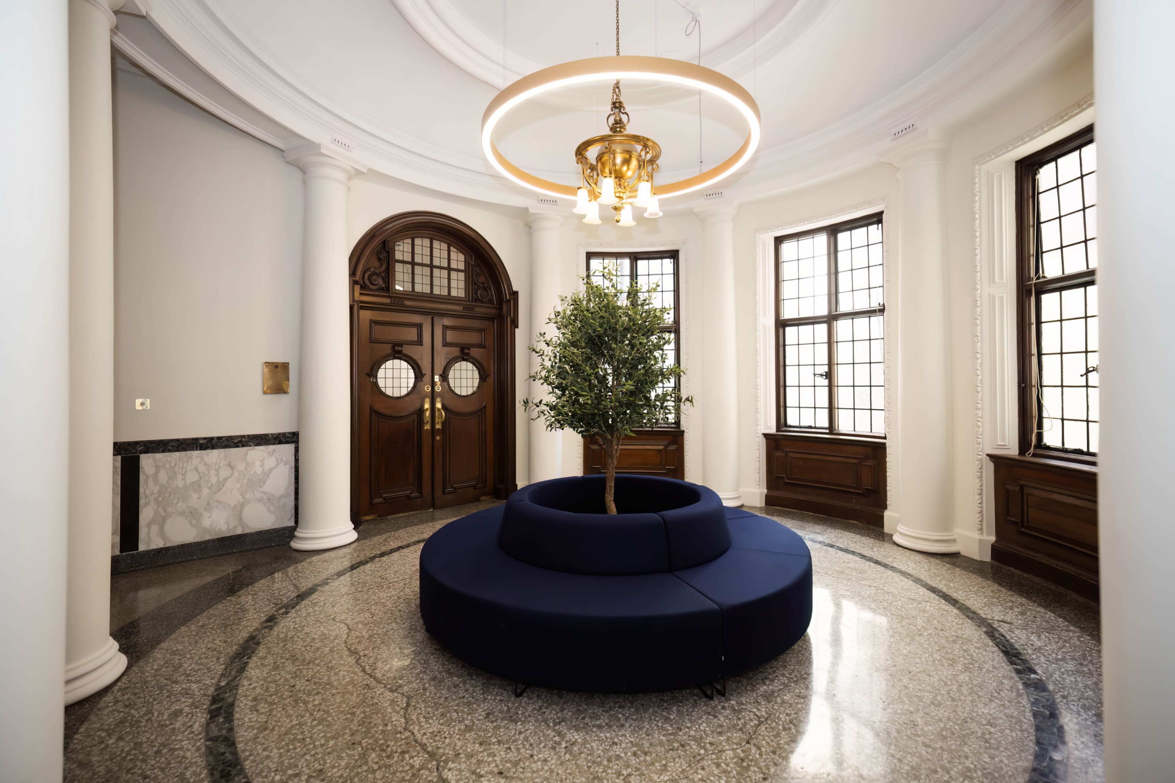 A circular seating area with a tufted navy blue sofa, placed in a well-lit entryway featuring large windows and wooden doors.