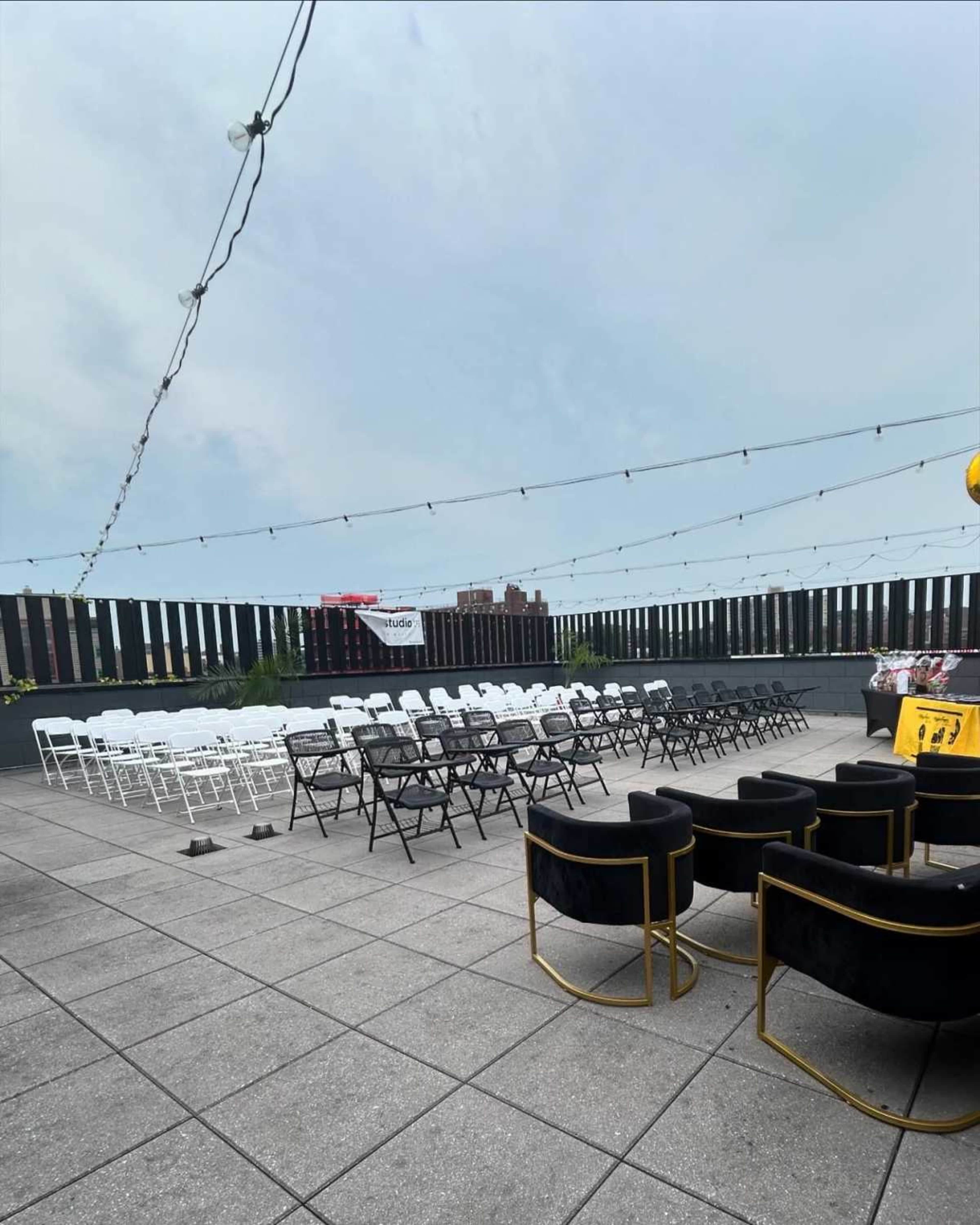 The image shows a rooftop area set up with rows of white and black chairs, and string lights overhead, against a backdrop of urban buildings.