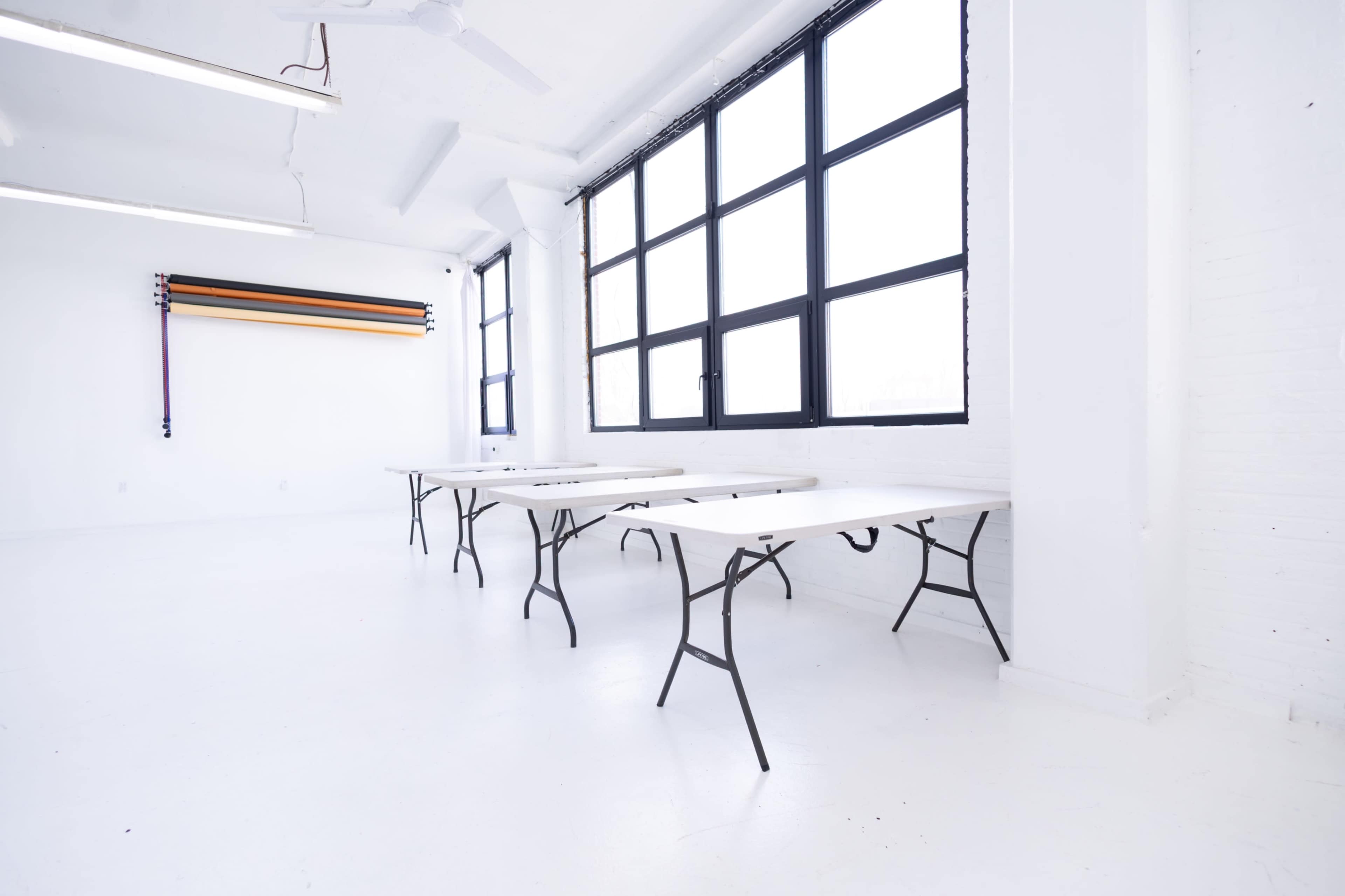 A bright, empty room with white walls and floors contains several long folding tables arranged parallel to large windows.
