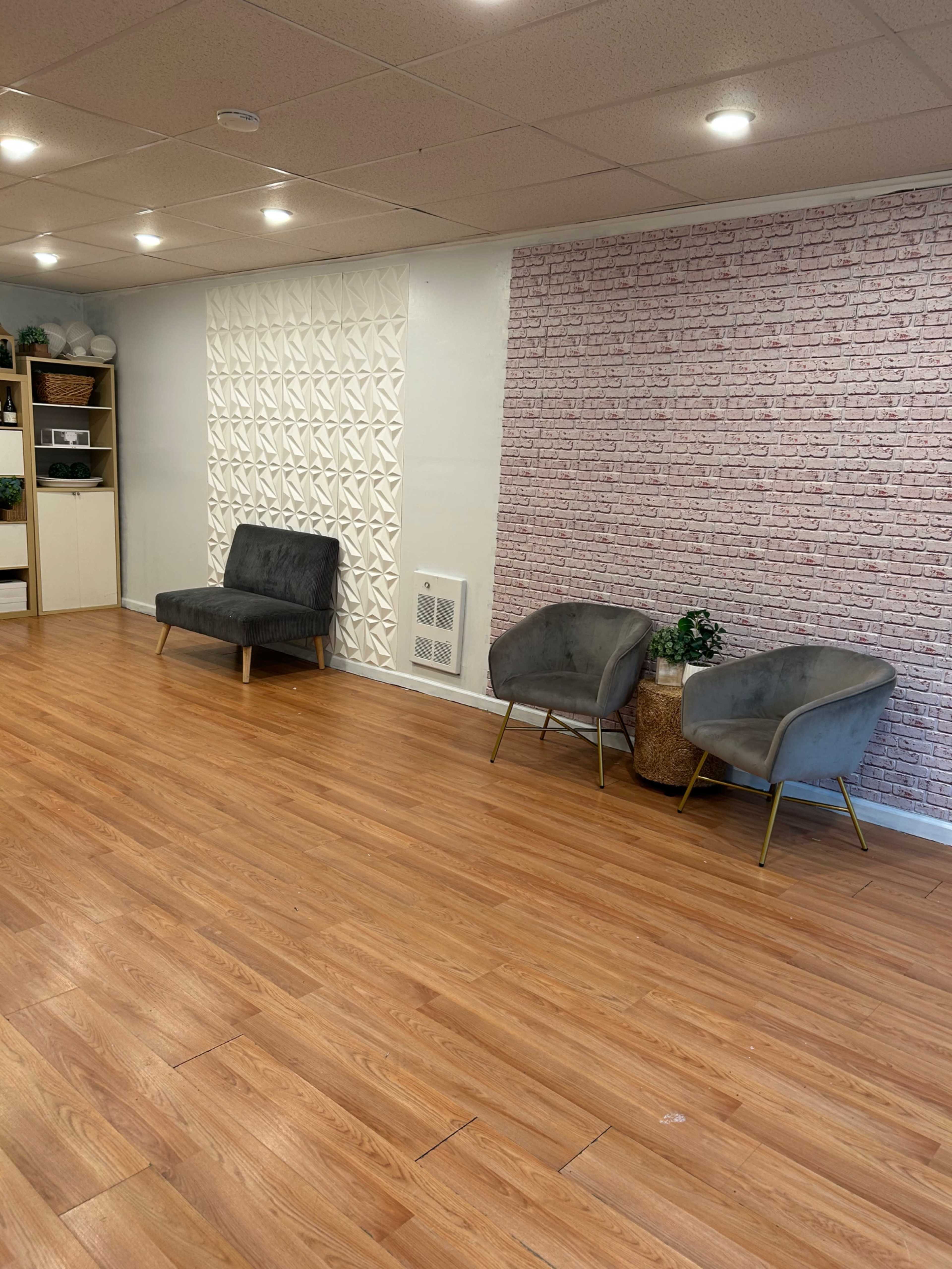 The image shows a spacious room with wooden flooring, featuring two gray chairs and a textured white and brick wall.