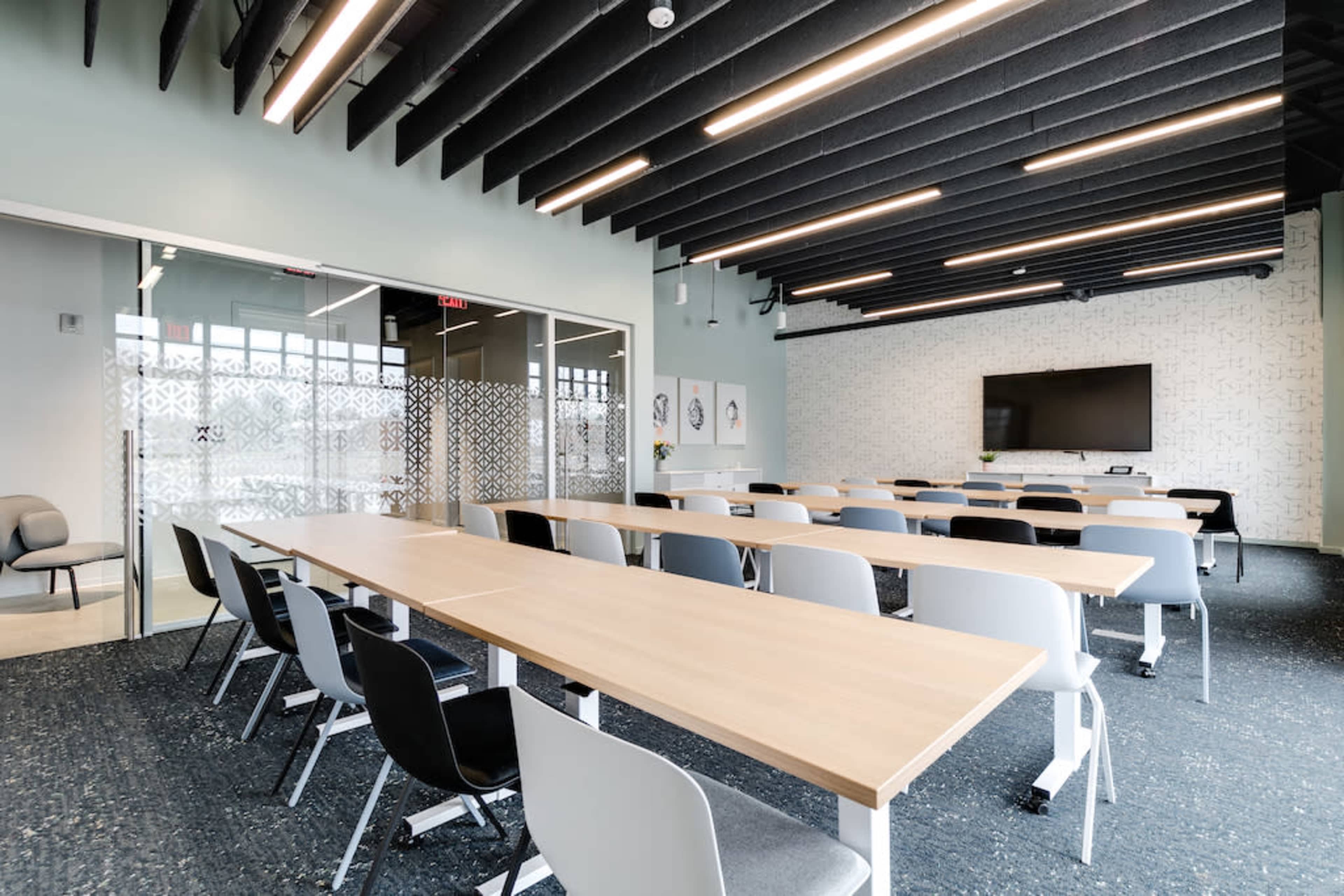 A modern conference room features long tables arranged in rows with chairs, large windows, and a wall-mounted screen.