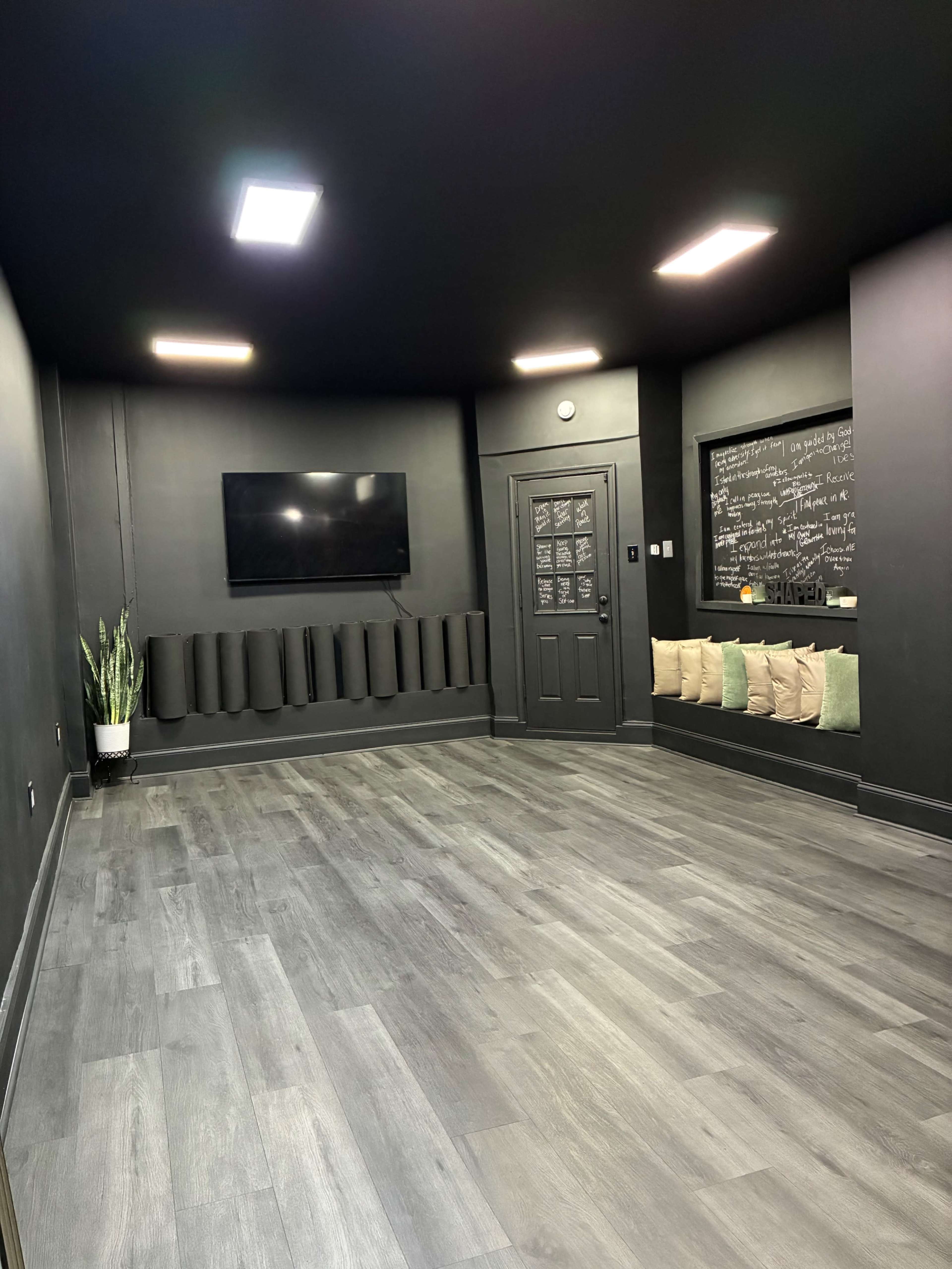 The image shows a modern, minimalist room with black walls, a flat-screen TV on one wall, a door with a window, and a seating area featuring cushions on a ledge.