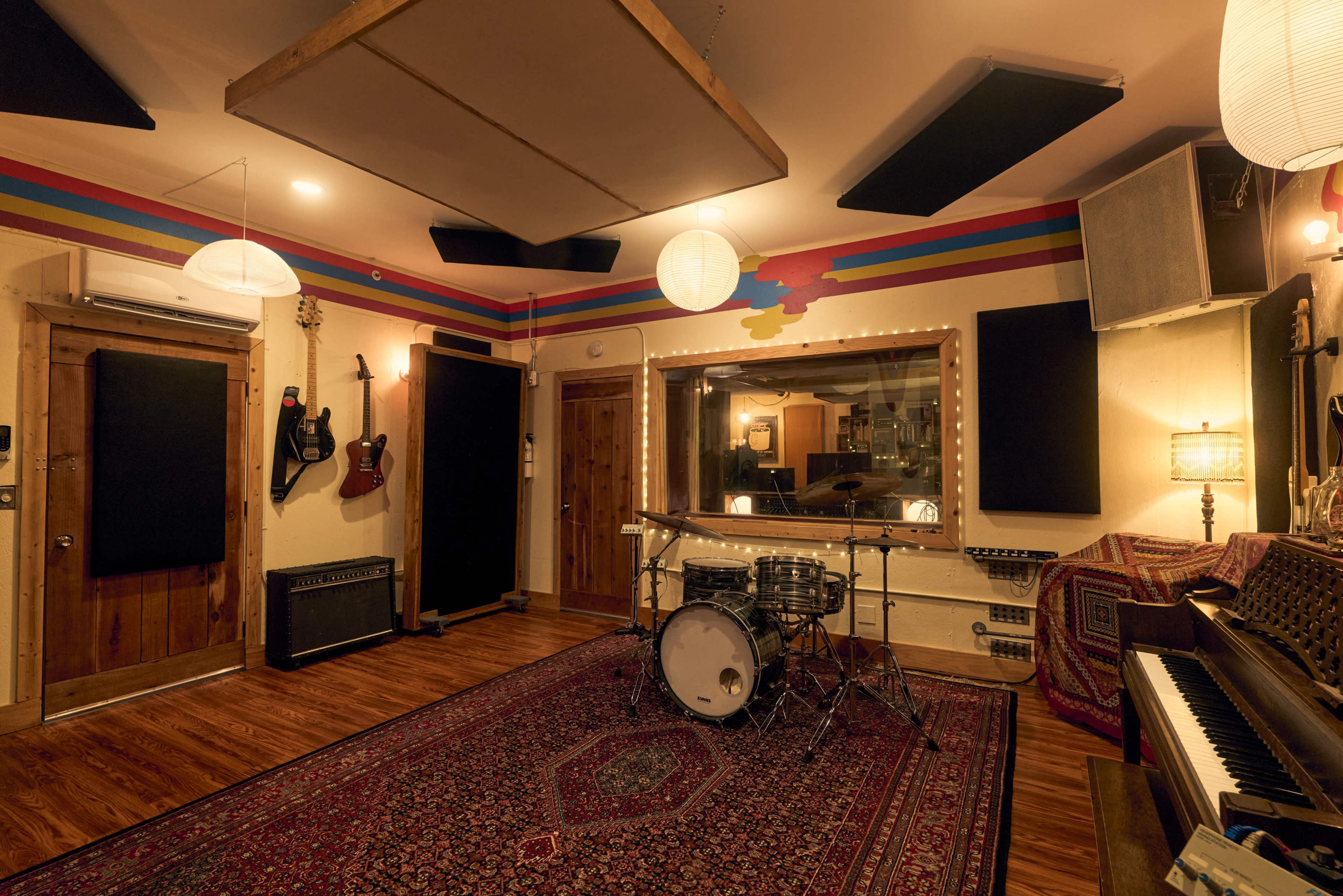 The image shows a music recording studio featuring a drum set, various musical instruments, acoustic panels on the walls, and warm lighting.