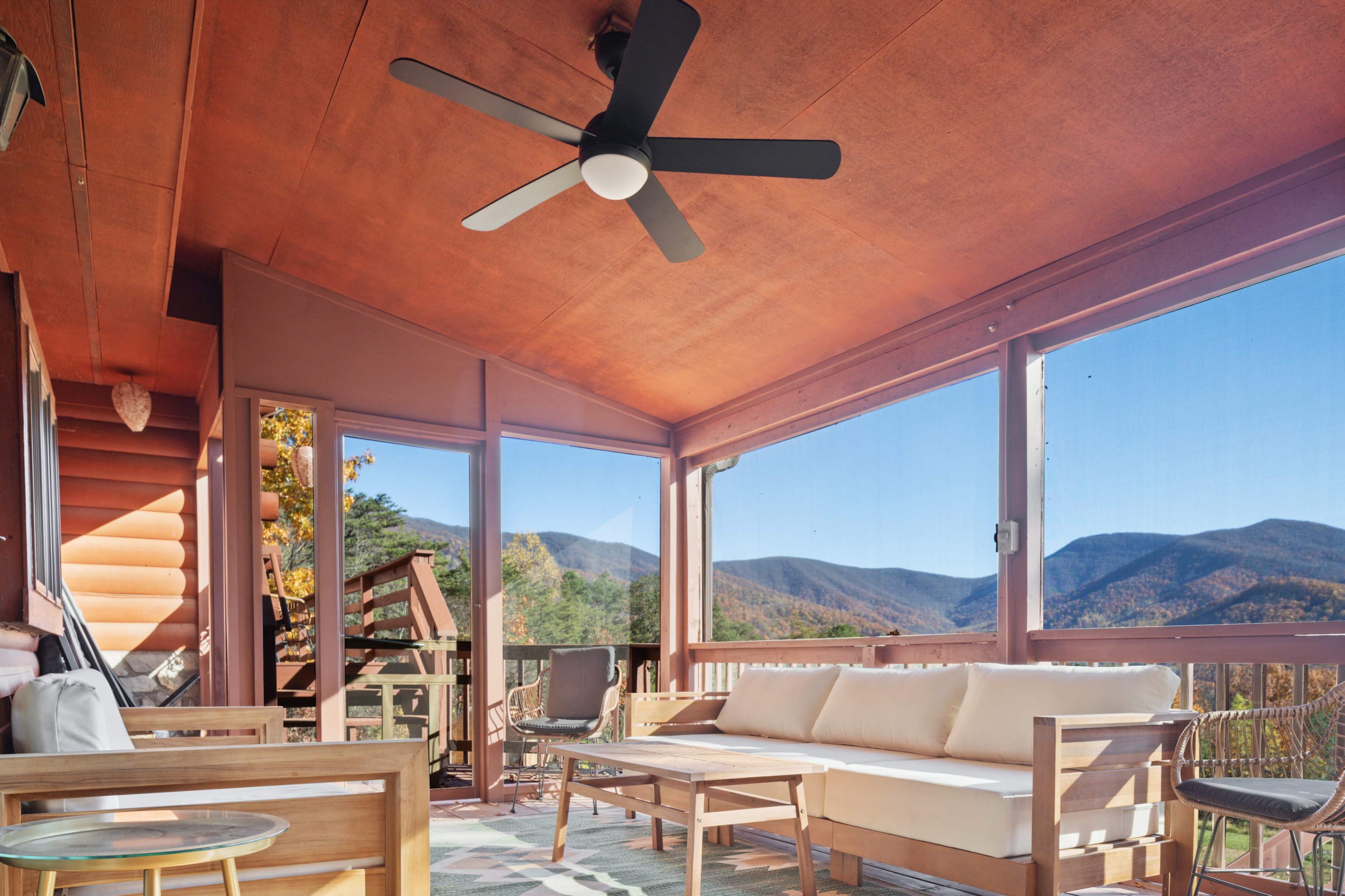 The image shows a screened-in porch with a sofa and a ceiling fan, overlooking scenic mountains in the background.