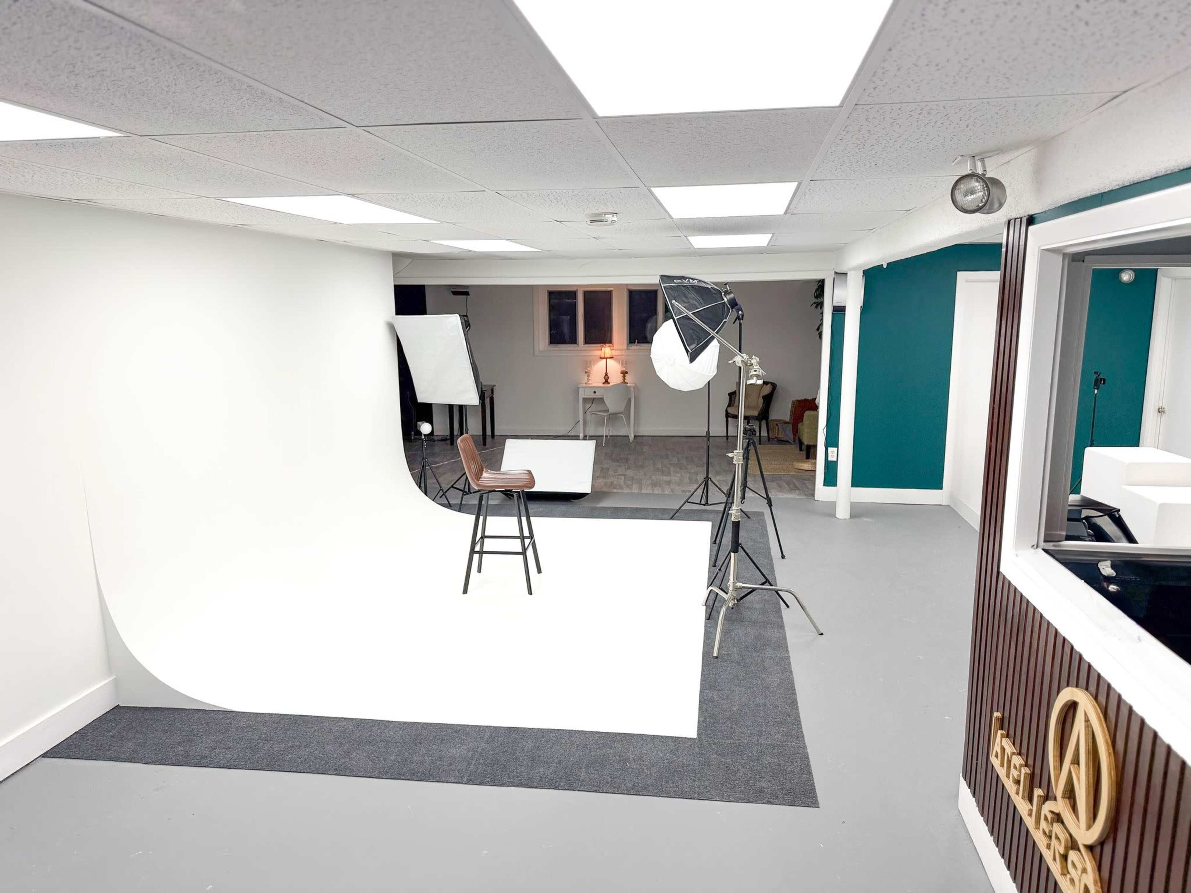 The image shows a photography studio with a white backdrop, a chair in front of a table, and various lighting equipment set up in a spacious room.