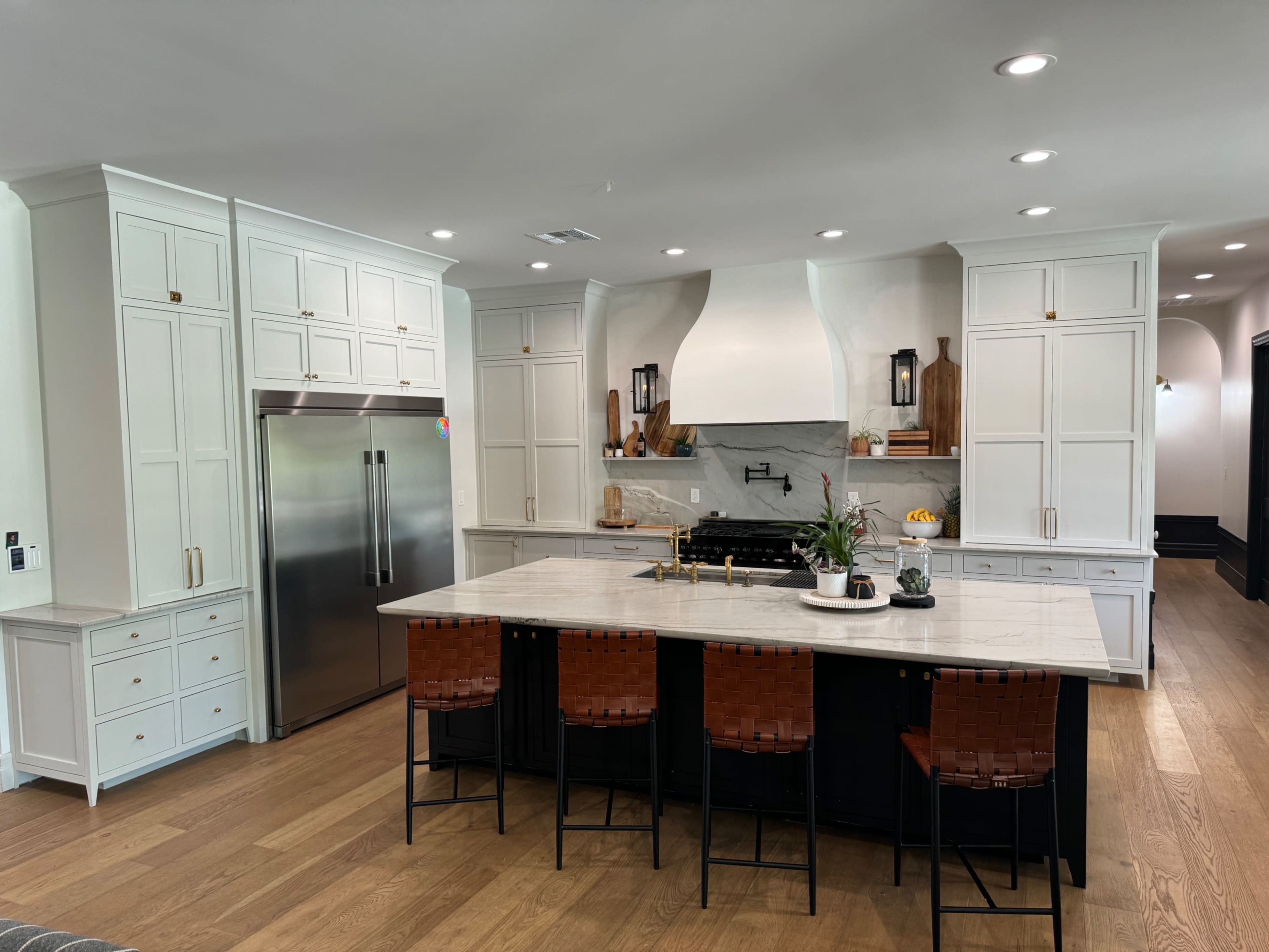 A modern kitchen featuring a central island with four bar stools, white cabinetry, a stainless steel refrigerator, and a marble countertop with a decorative arrangement on top.