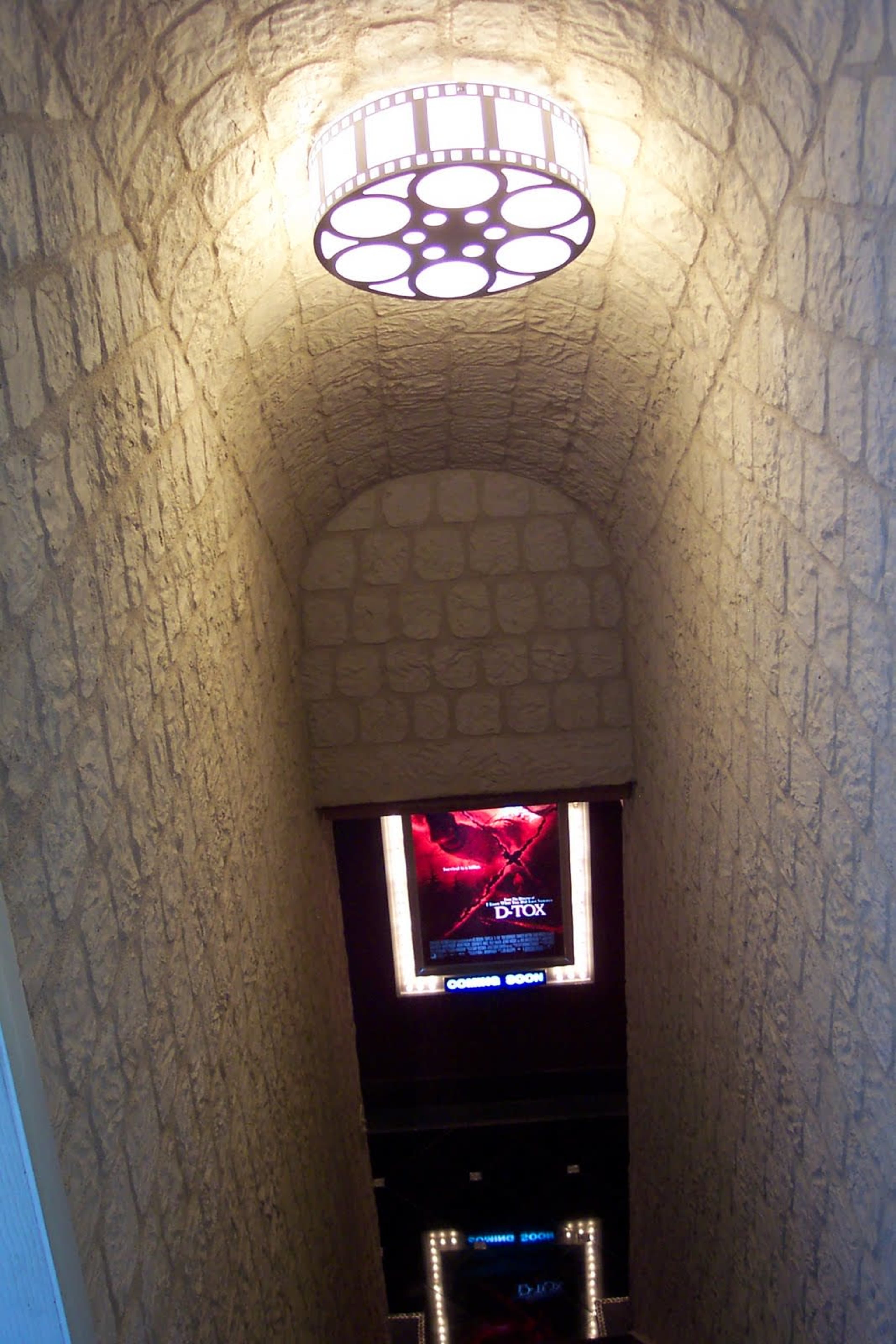 A stone-walled stairway leads down to a lit area featuring a movie poster and a circular light fixture resembling a film reel.