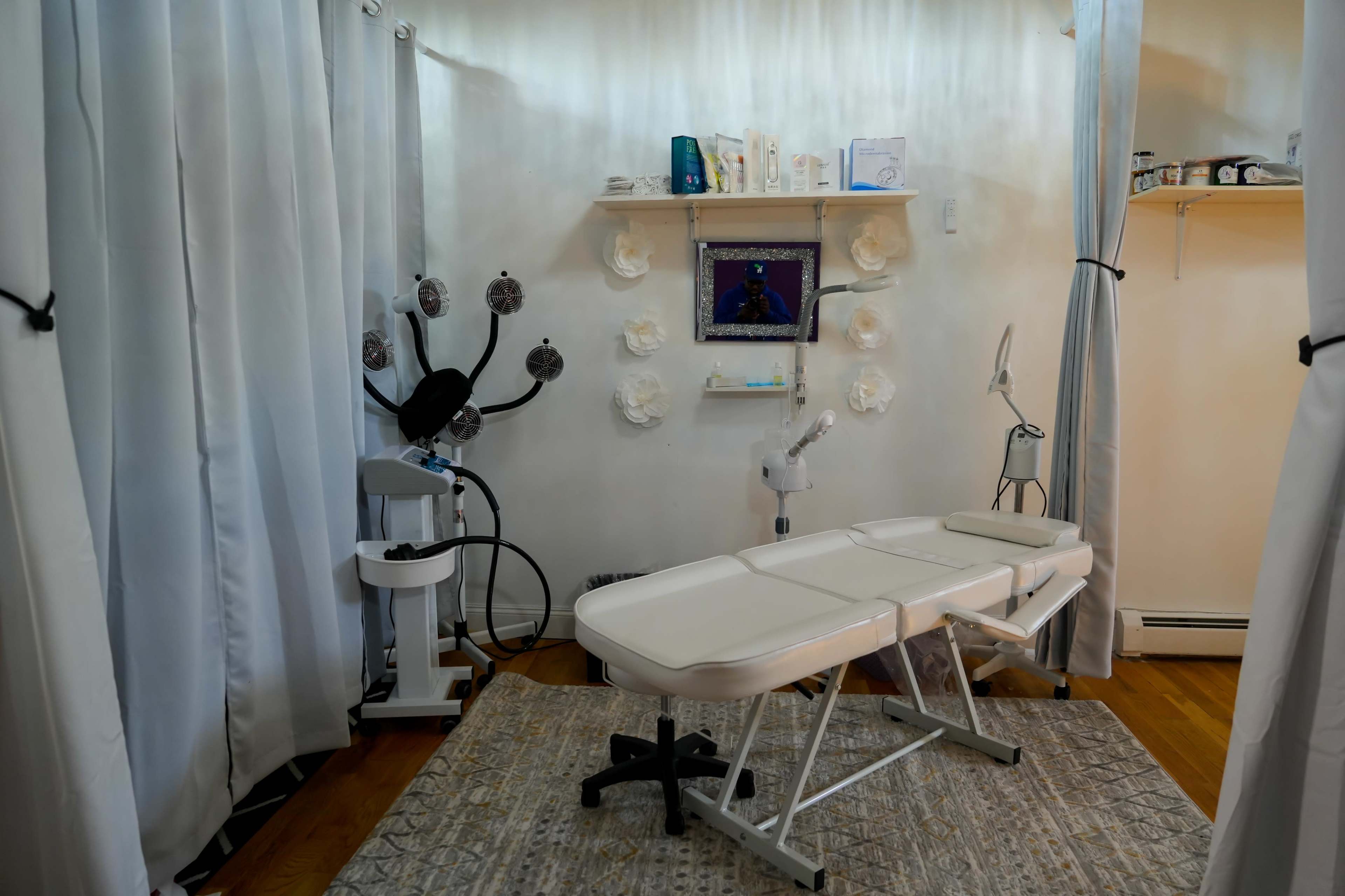 A treatment room with a massage table, a facial steamer, a shelf displaying beauty products, and white curtains separating the space.