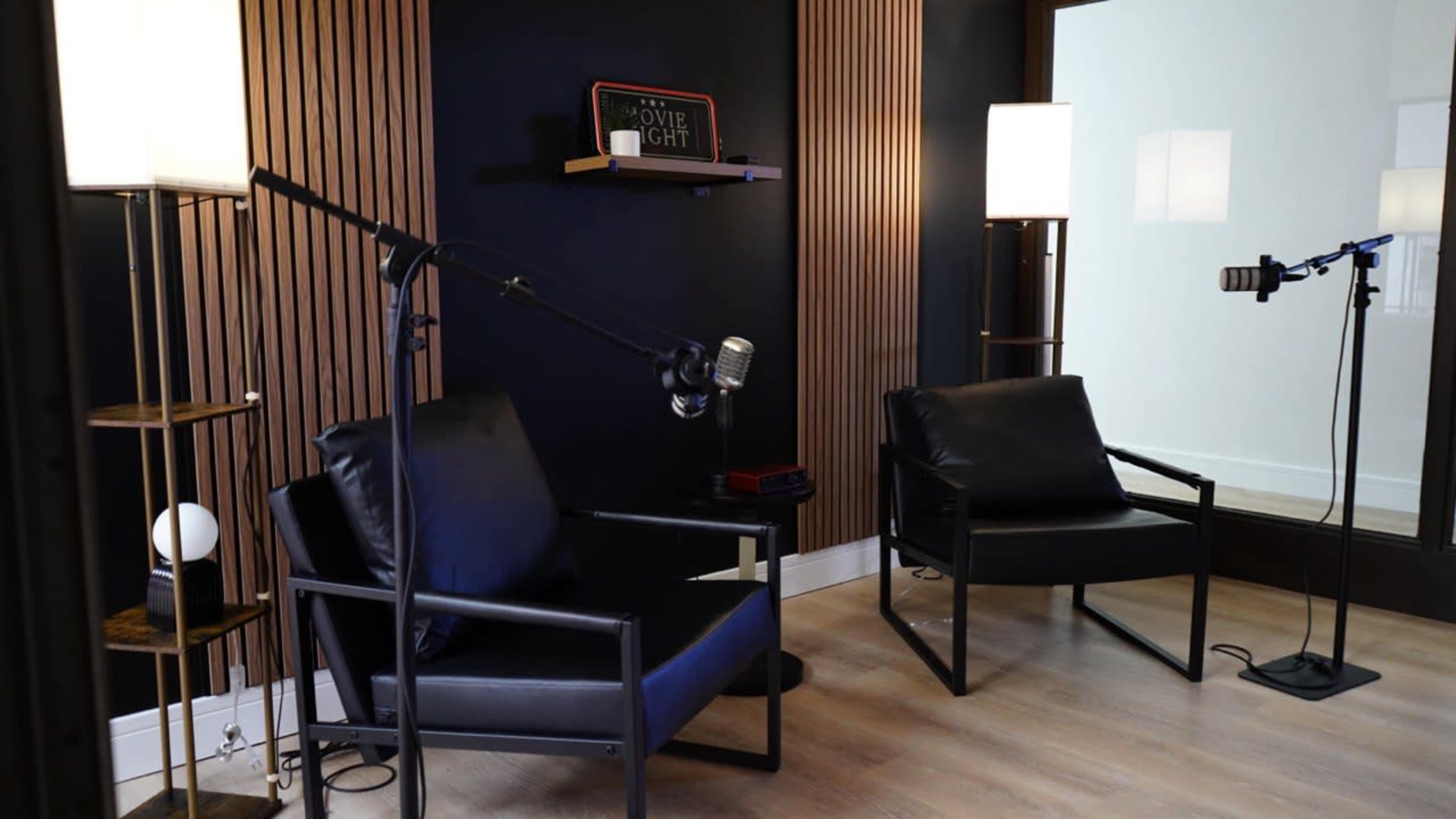 The image shows a cozy interview setup featuring two black armchairs, a microphone on a stand, and soft lighting against a dark wall with wooden slats.