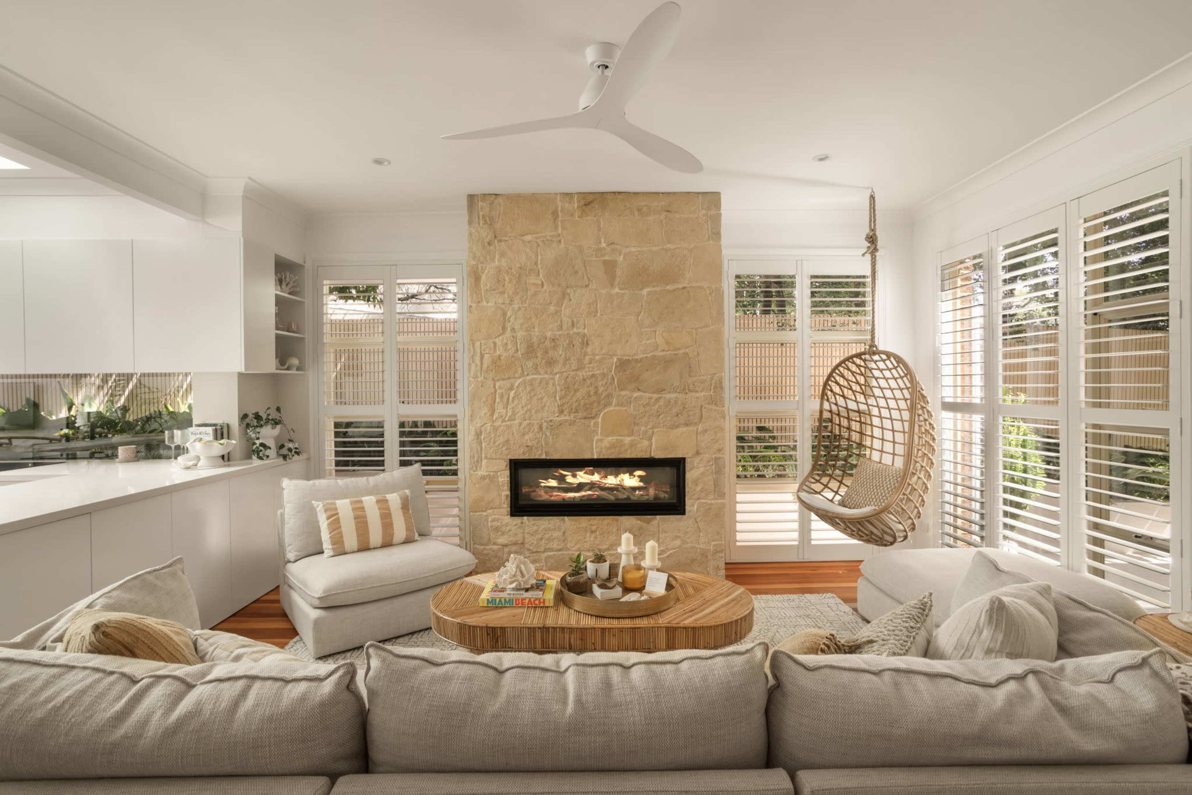 The image features a cozy living room with a stone fireplace, a large sectional sofa with plush cushions, and a hanging chair near windows with plantation shutters.