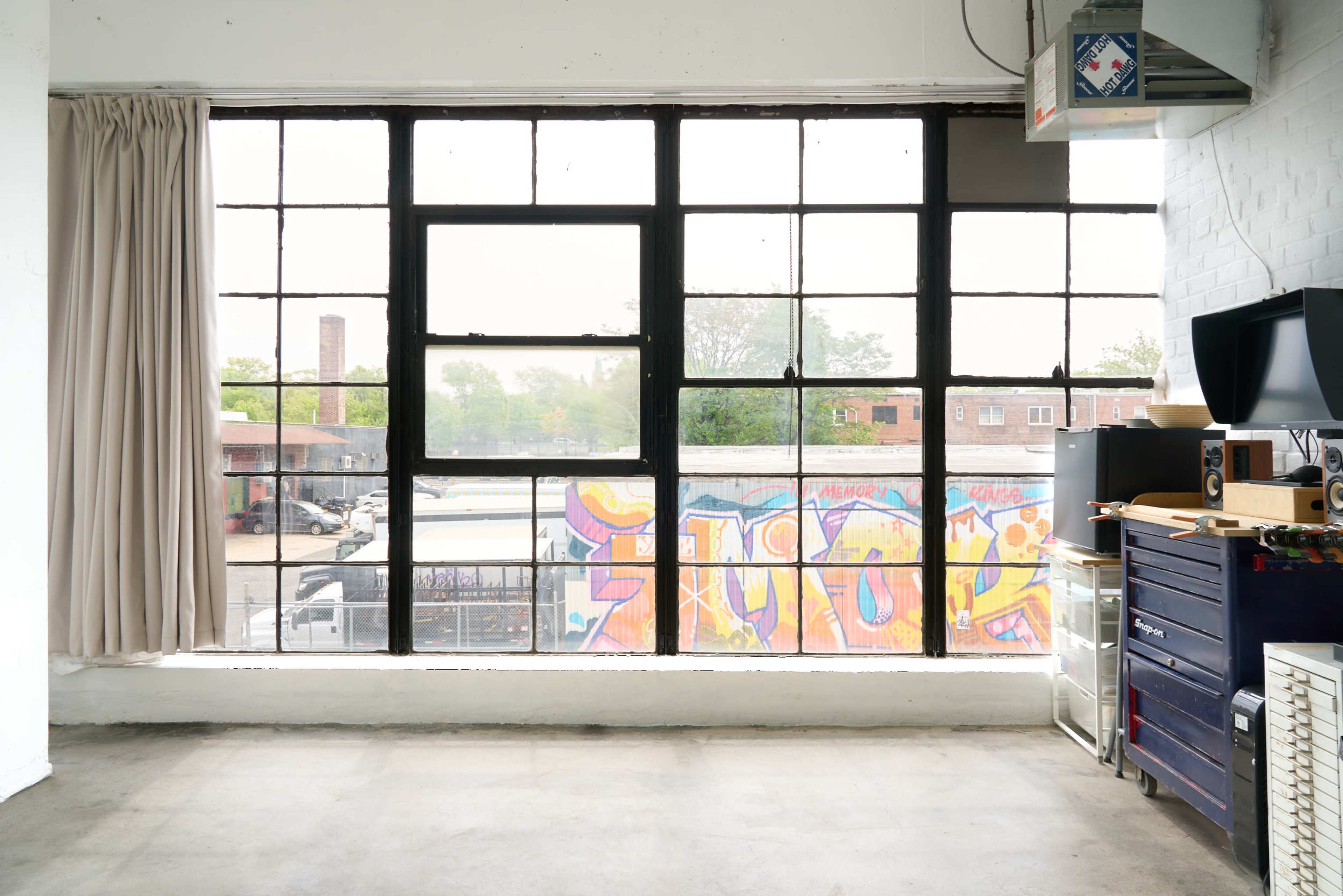 The image shows a large industrial window with black frames overlooking a parking area, featuring colorful graffiti on the exterior wall.