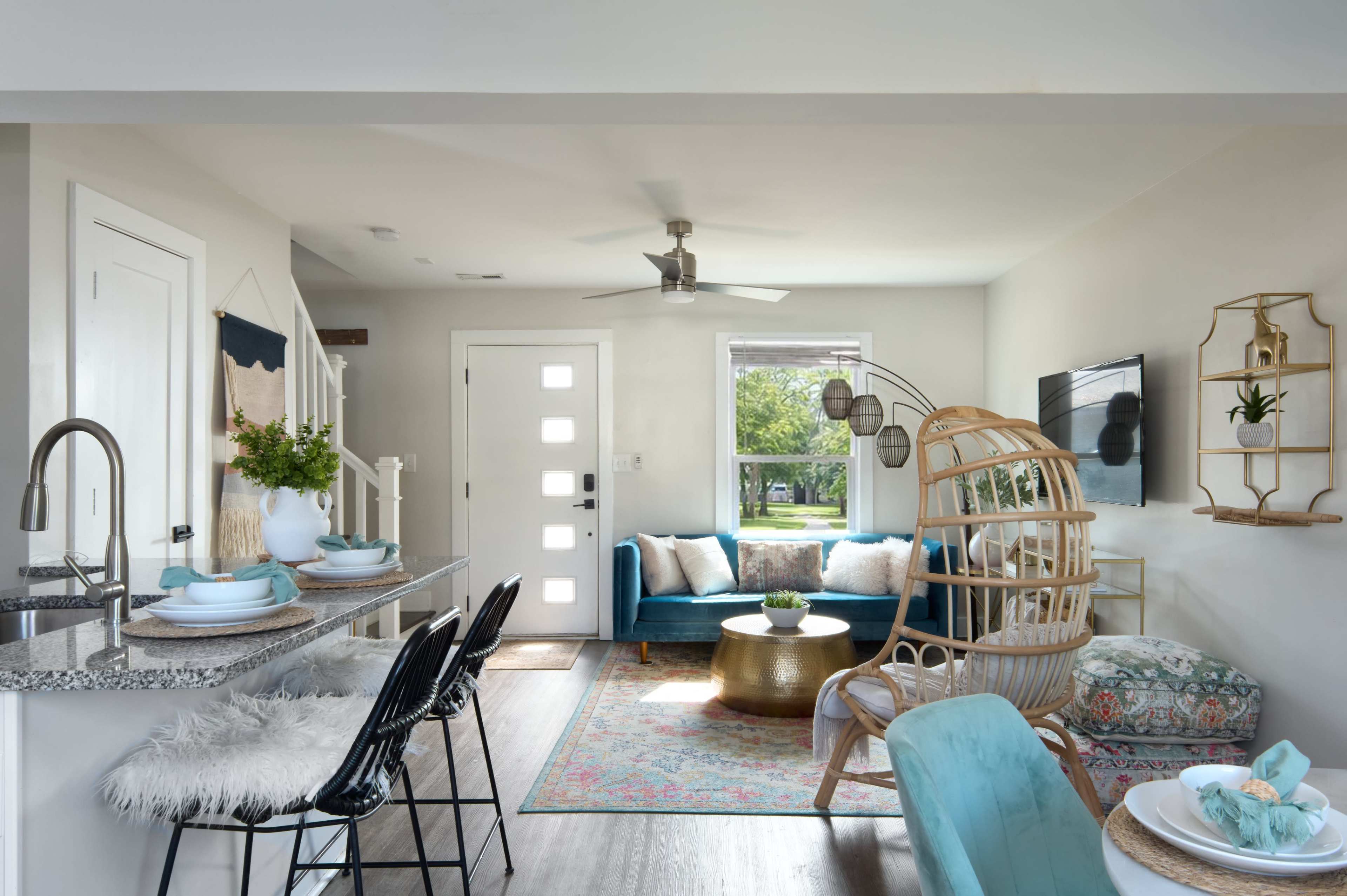 The image shows a modern open-concept living space featuring a kitchen island with bar stools, a cozy seating area with a blue couch, and a birdcage-style chair, along with a dining table set for meals.