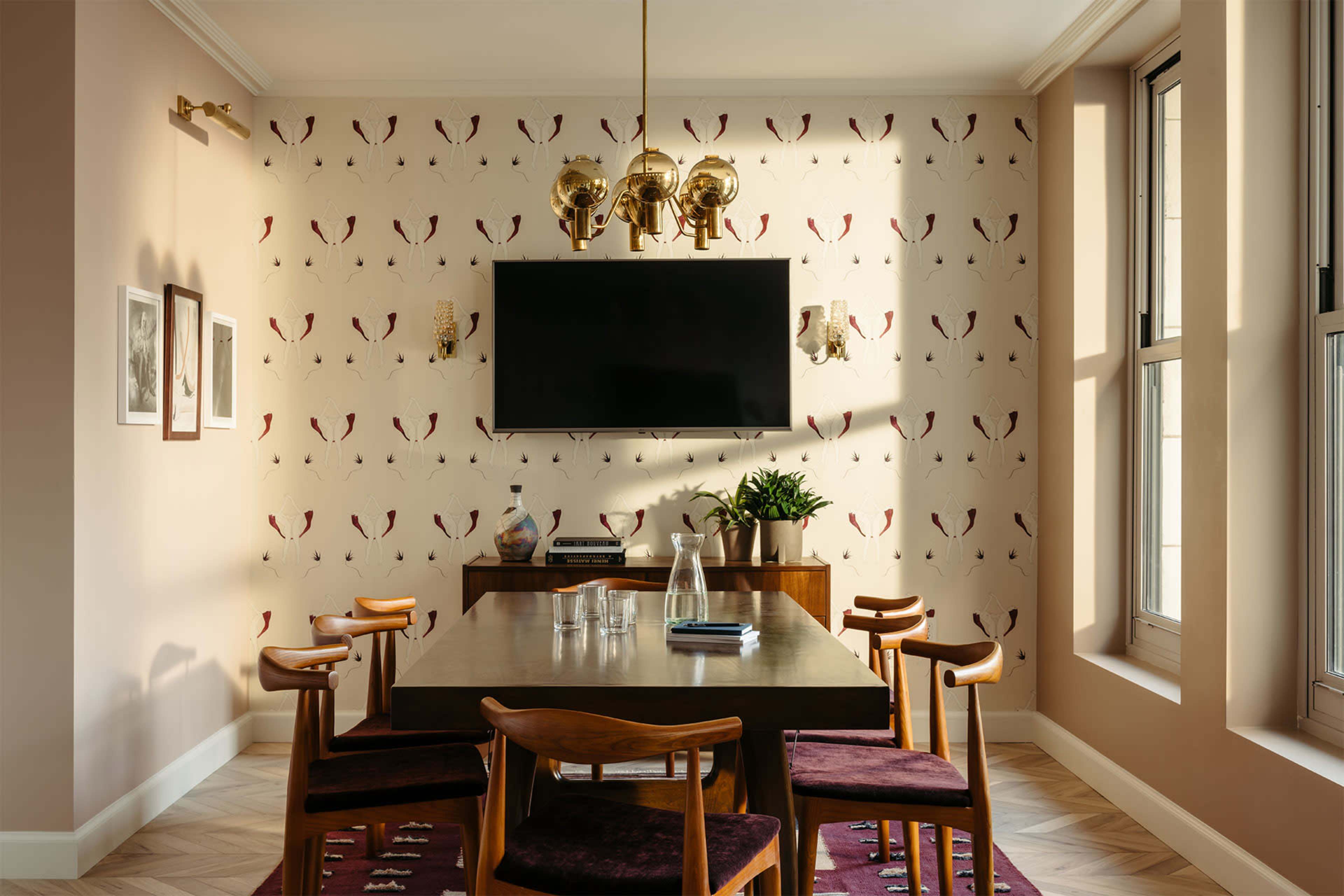 A dining room features a wooden table surrounded by chairs, with a wall adorned in patterned wallpaper and a mounted television.
