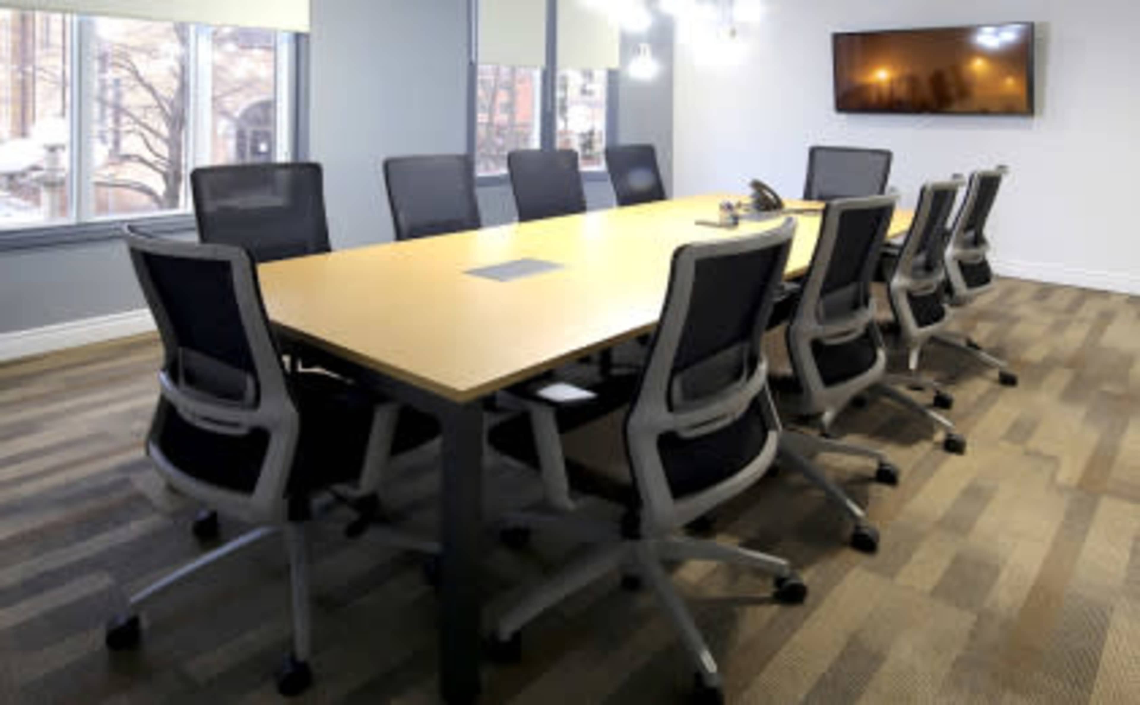 A long conference table surrounded by ergonomic chairs is set up in a well-lit meeting room with large windows and a wall-mounted screen.