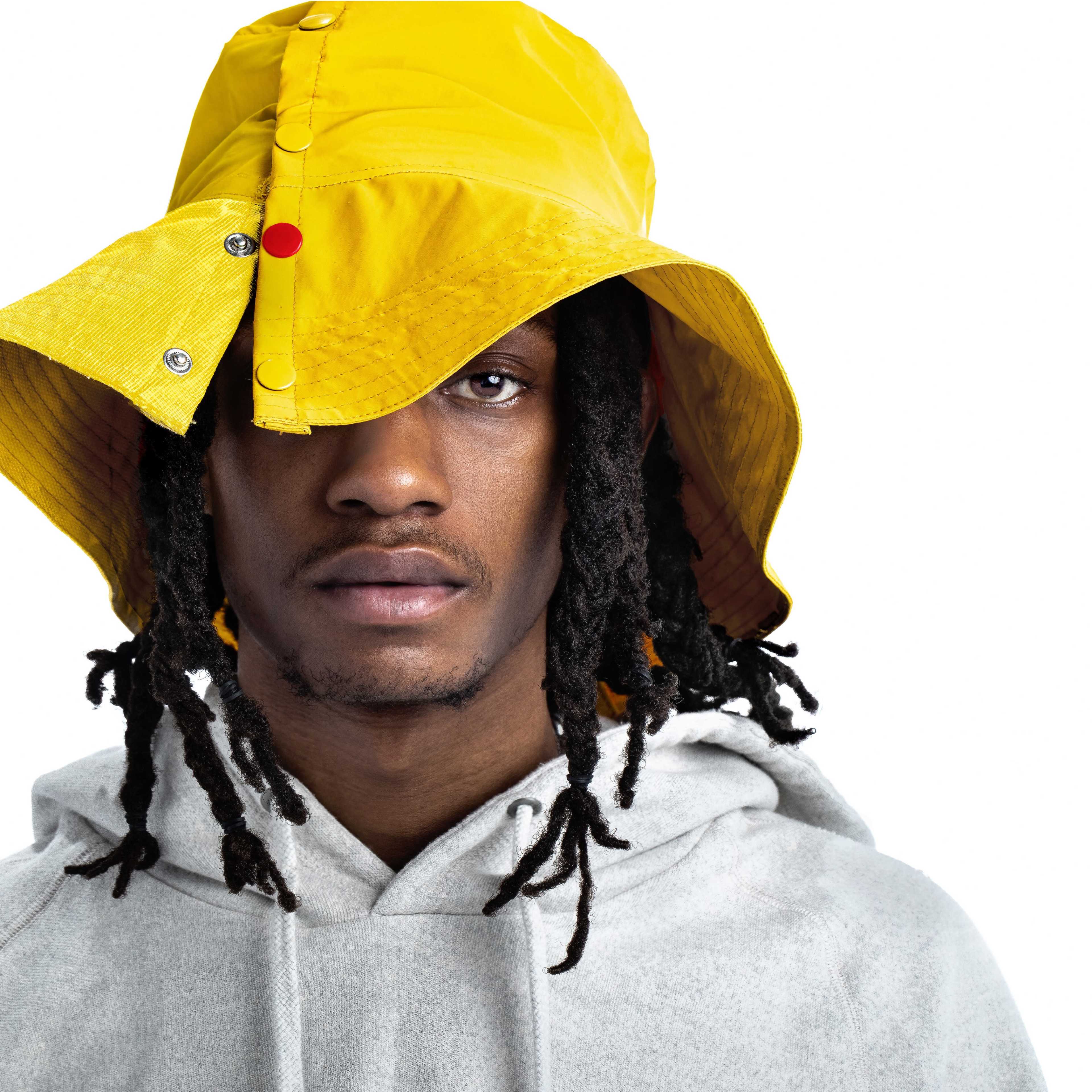 A young man wearing a yellow bucket hat and a gray hoodie looks directly at the camera.
