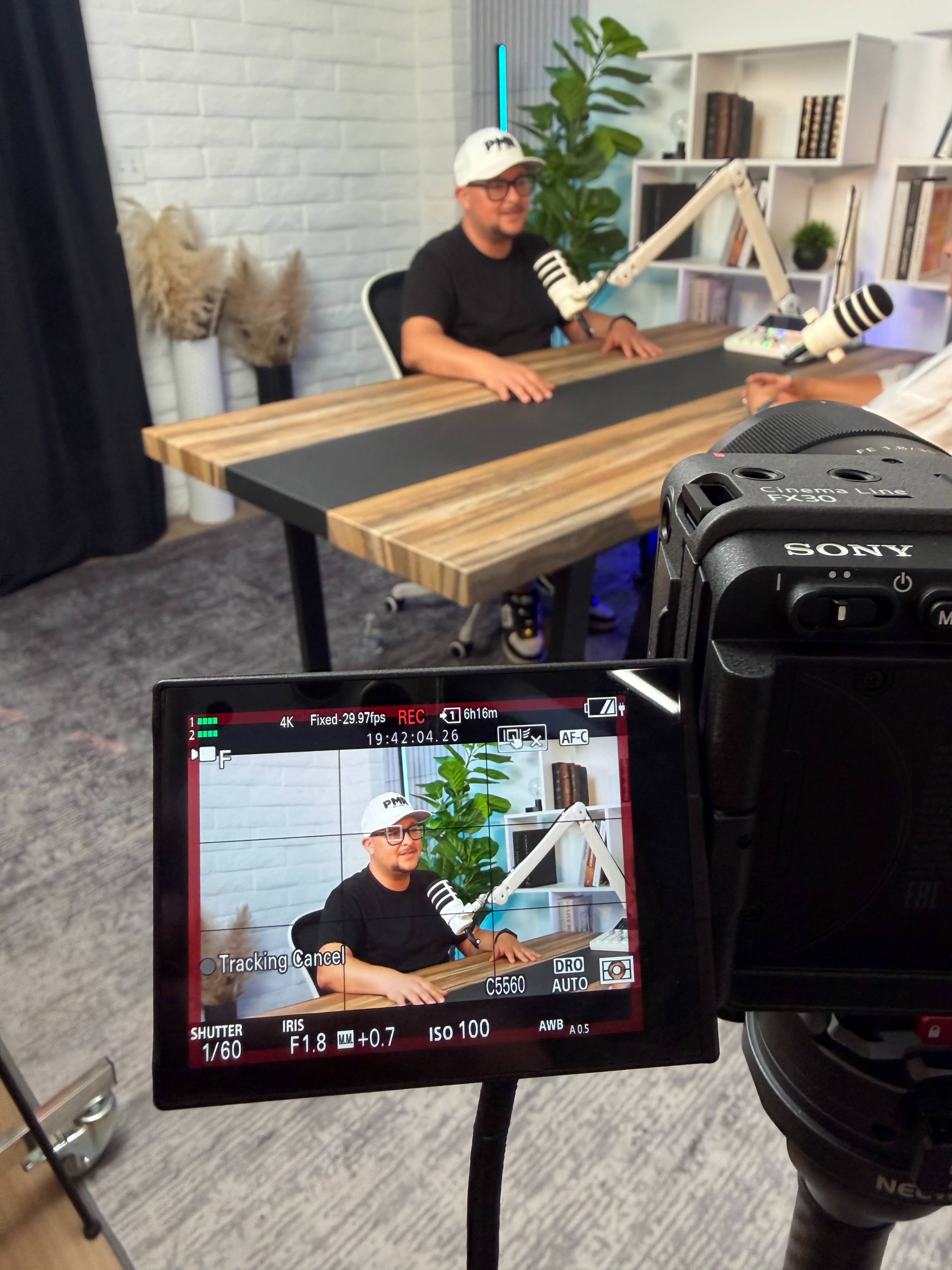 A person wearing a cap sits at a wooden desk in a well-lit studio setup, with a camera focused on them from a distance.