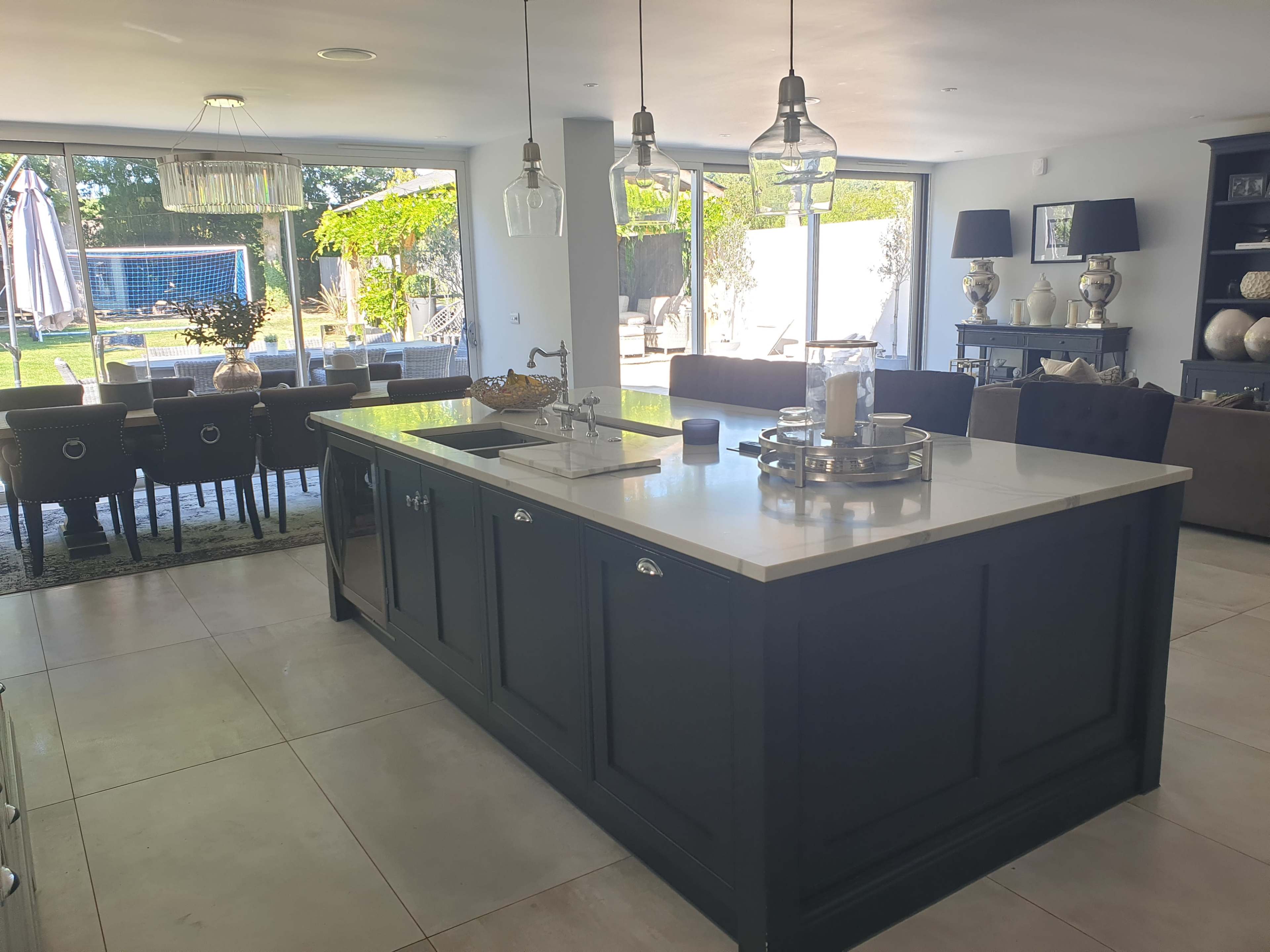 A modern kitchen with a large dark island, a dining area in the background, and large windows overlooking a garden.