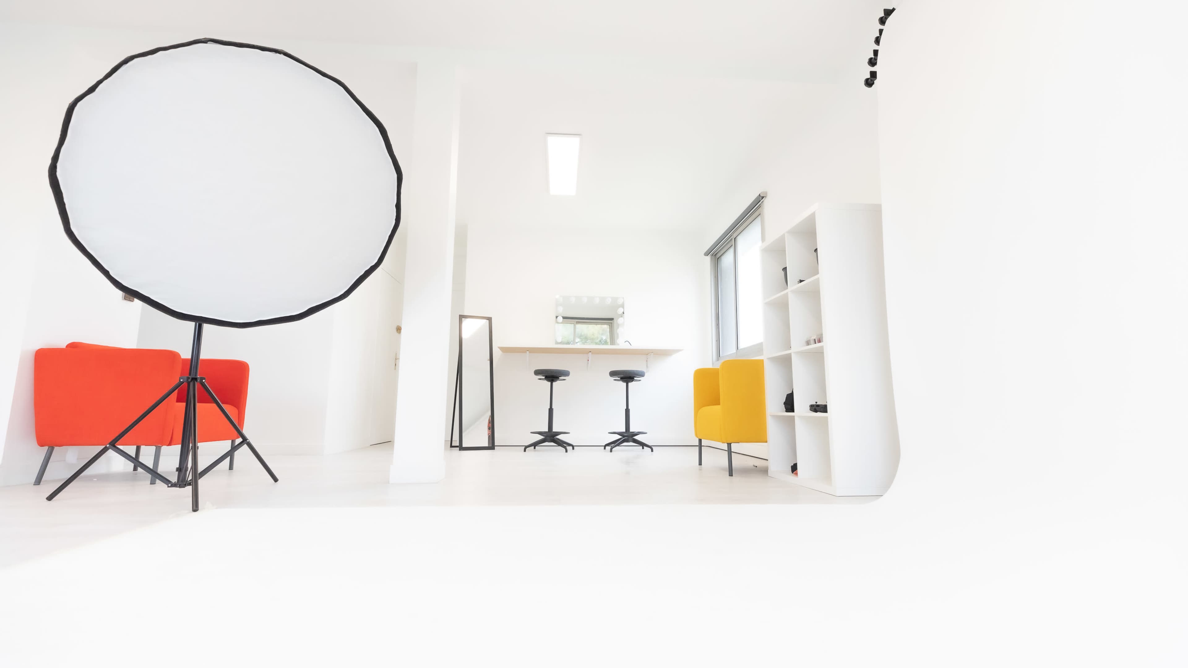 A bright, minimalist studio features a large reflector, two colorful chairs, a mirror, and a shelf against white walls.