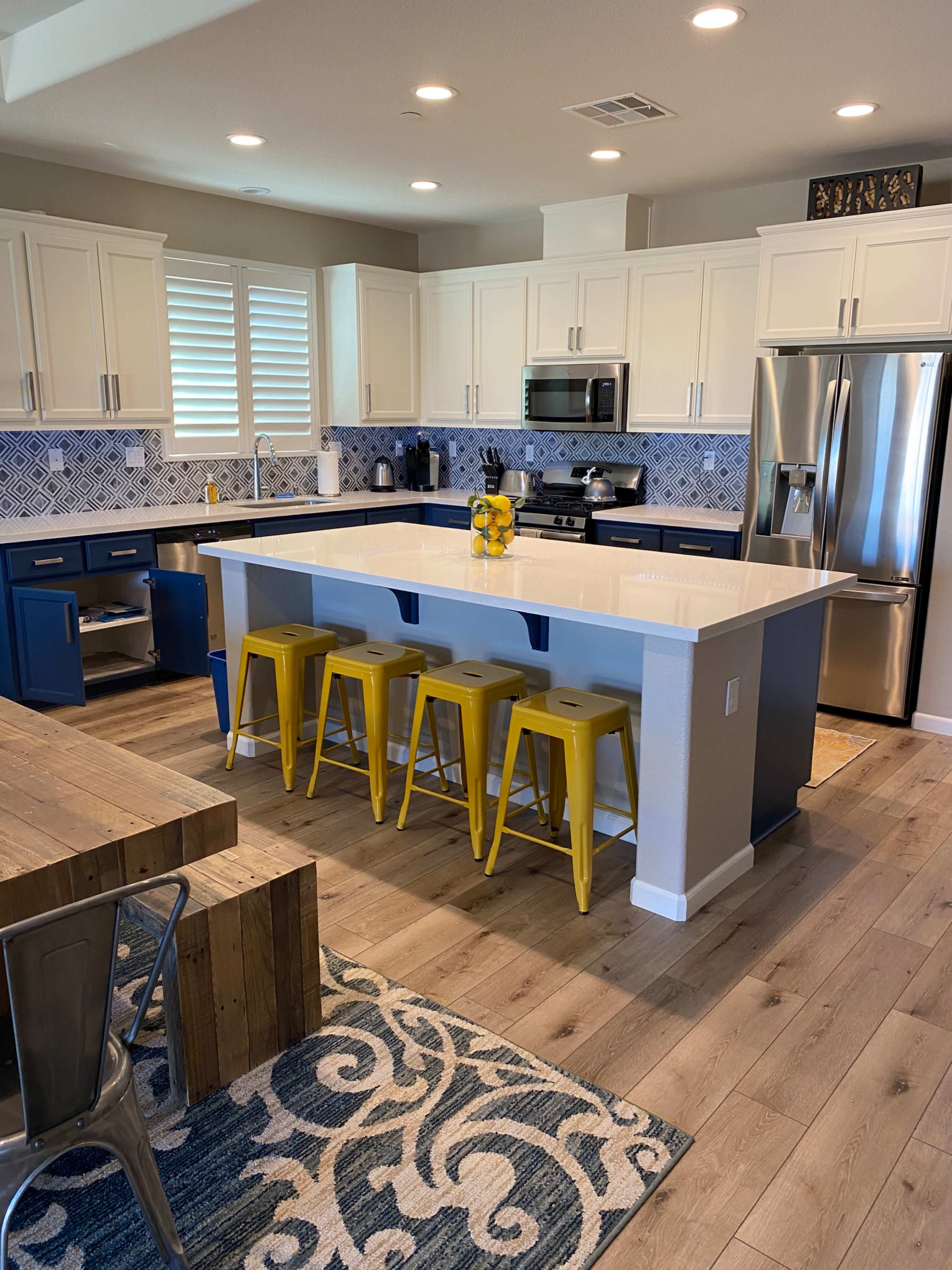 A modern kitchen features white cabinets, a blue island with yellow stools, stainless steel appliances, and a patterned rug.