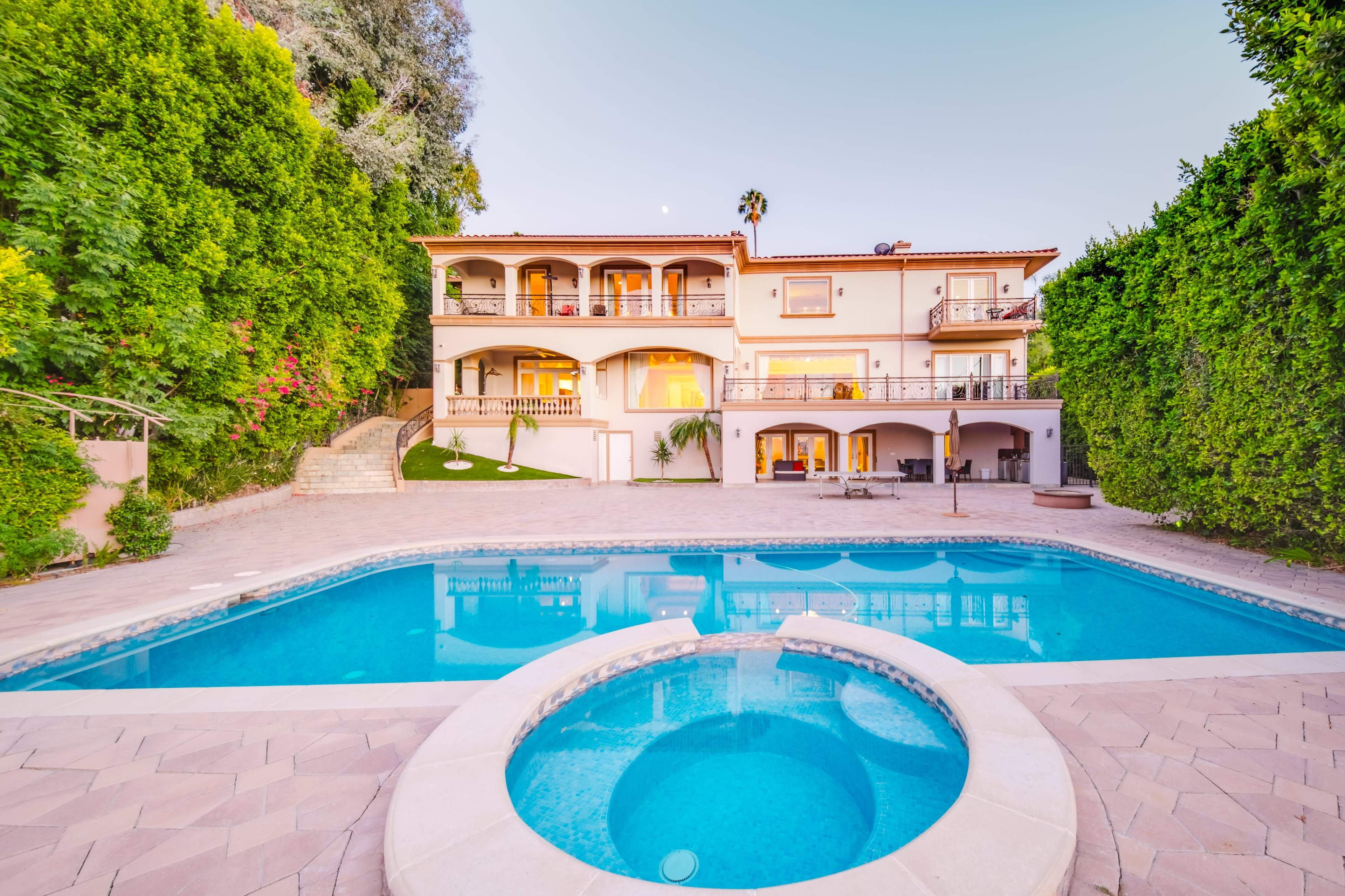 The image shows a large hillside house with a pool and jacuzzi, surrounded by lush greenery.