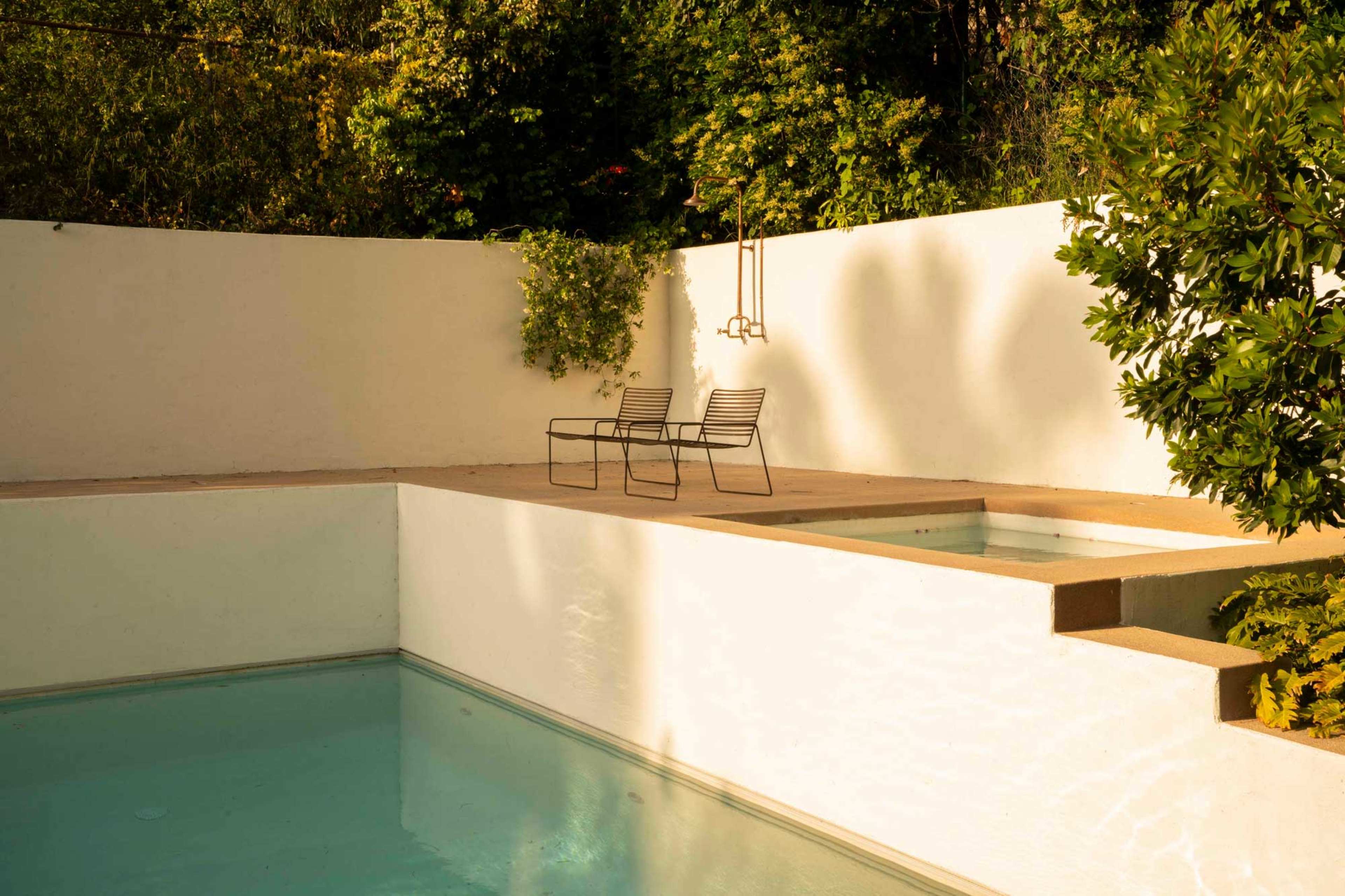 The image shows a pool area featuring two black metal chairs beside a bordered swimming pool with clear water and a white wall surrounded by greenery.